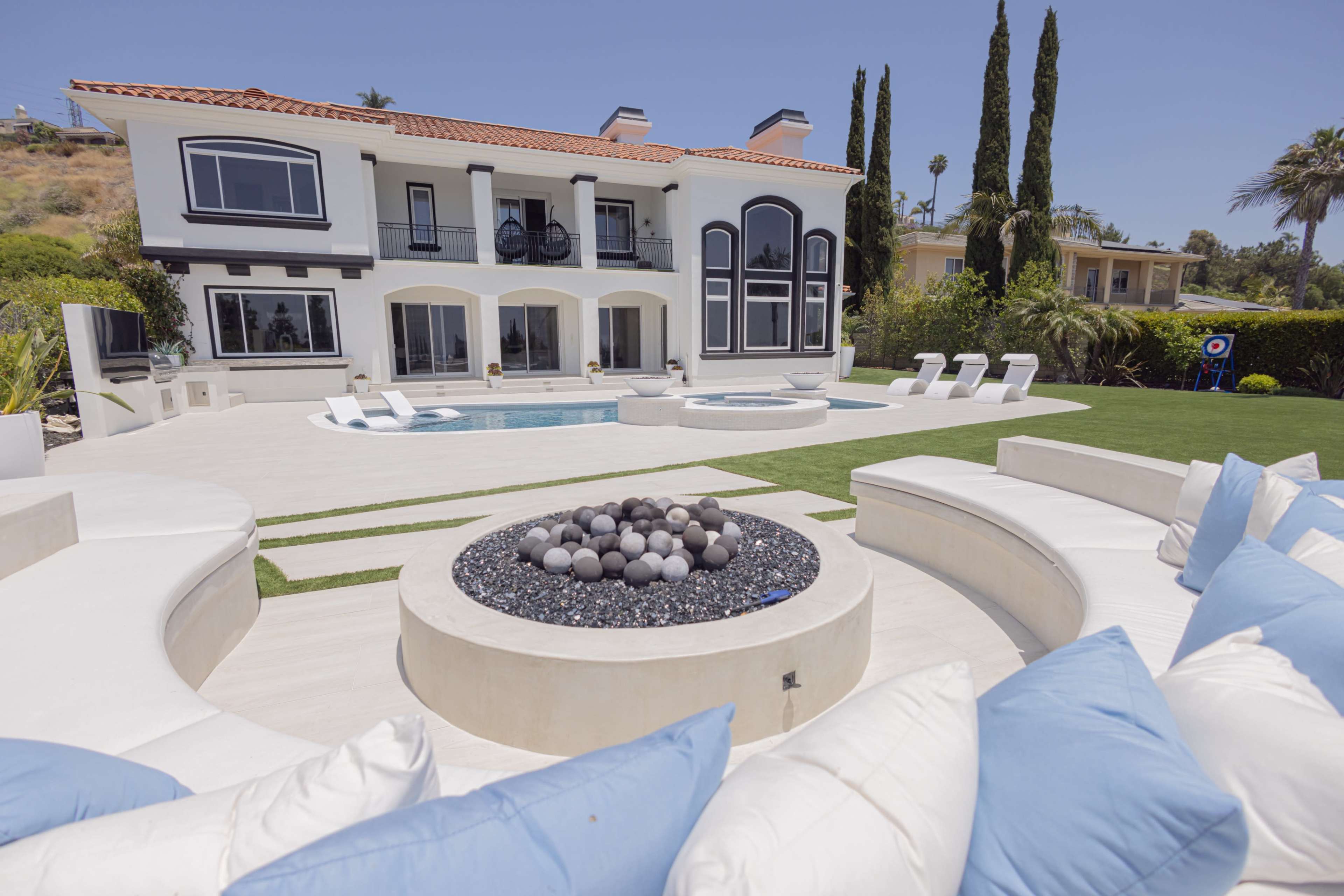 A luxurious outdoor area featuring a modern two-story villa, a swimming pool, and a circular seating area with a fire pit.