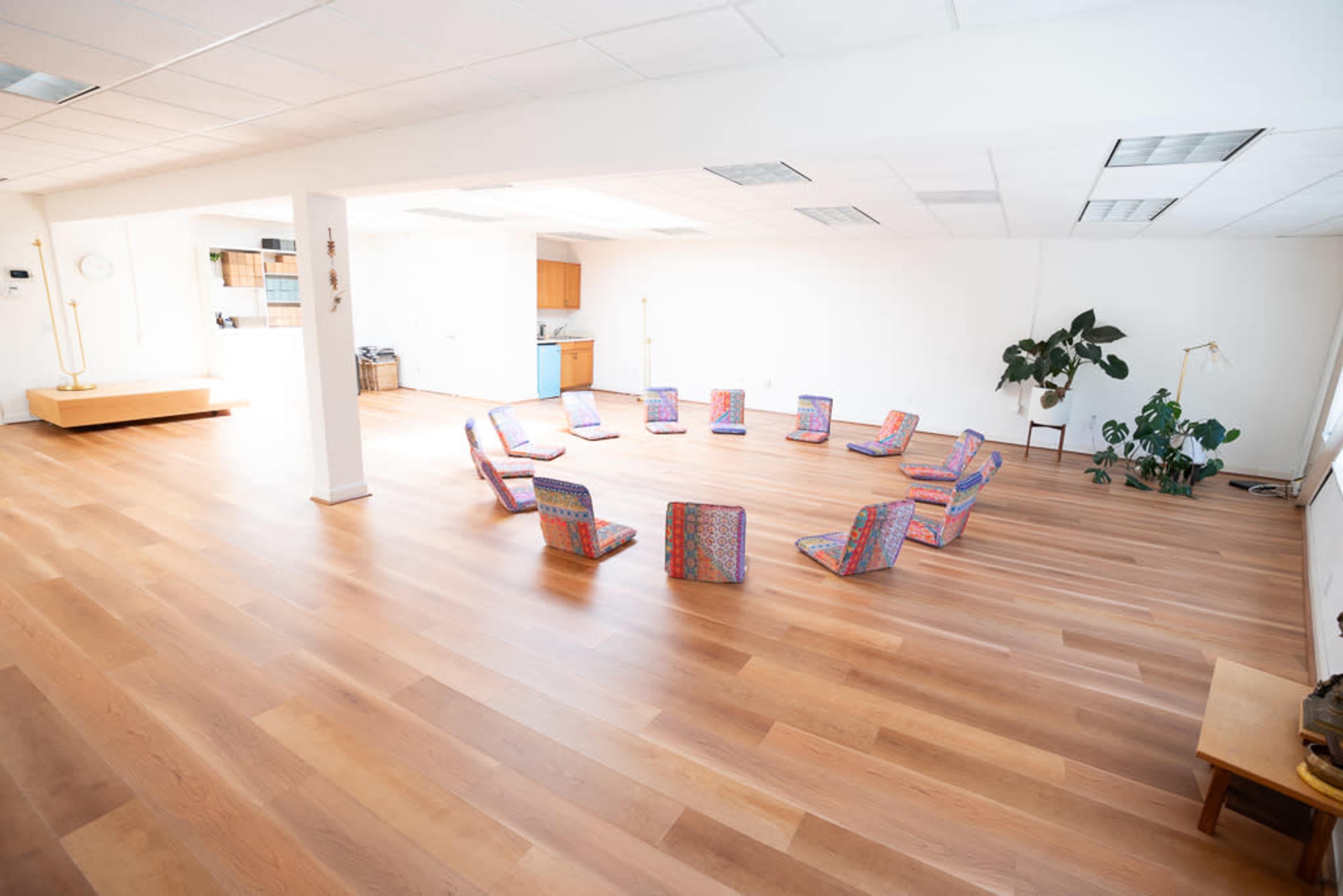 A spacious room with wooden flooring, arranged with colorful chairs in a circular formation and a small kitchenette visible in the background.