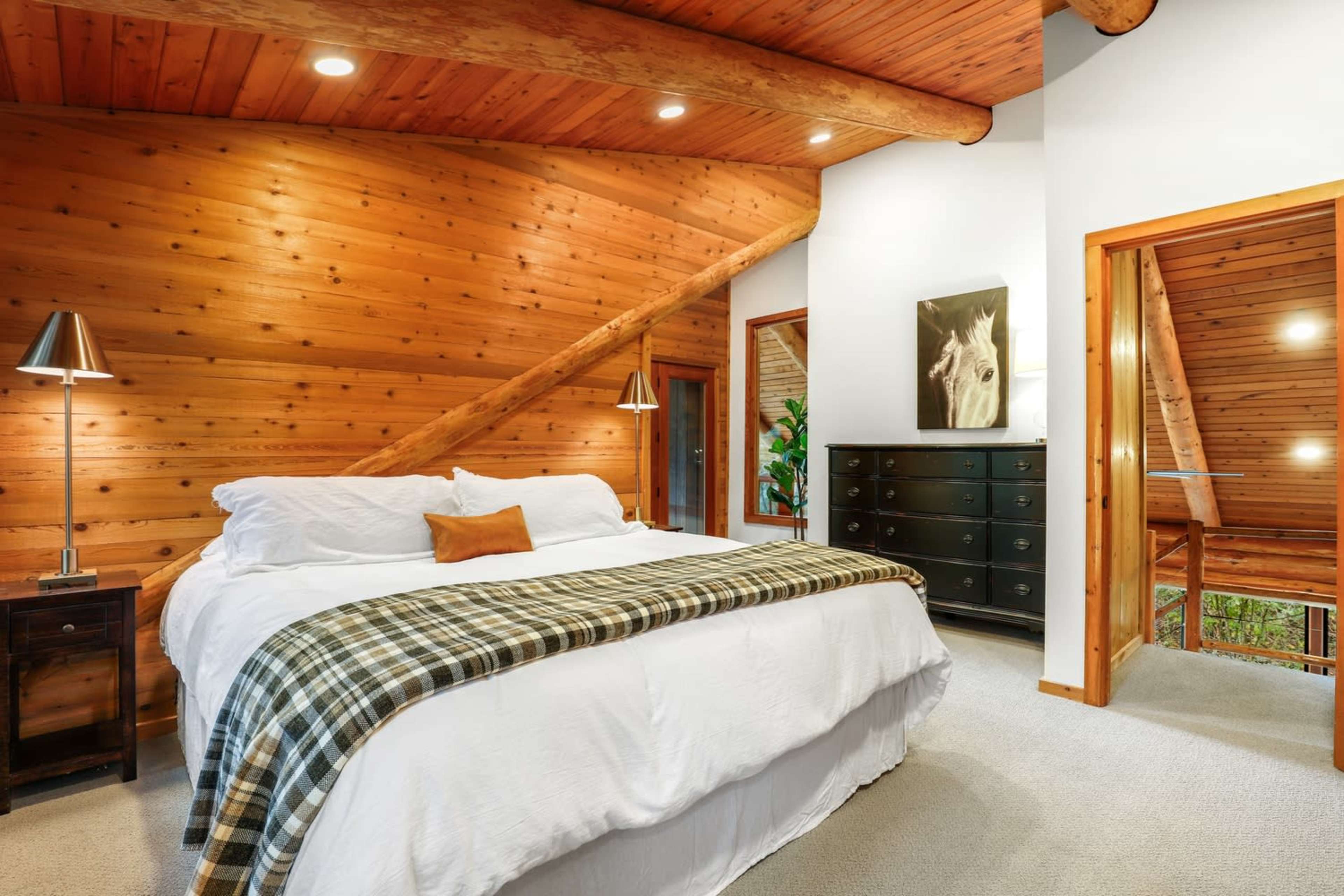 The image shows a cozy bedroom with wooden walls, a large bed dressed in white linens, and a decorative throw blanket, alongside a dark chest of drawers and a lamp on a nightstand.