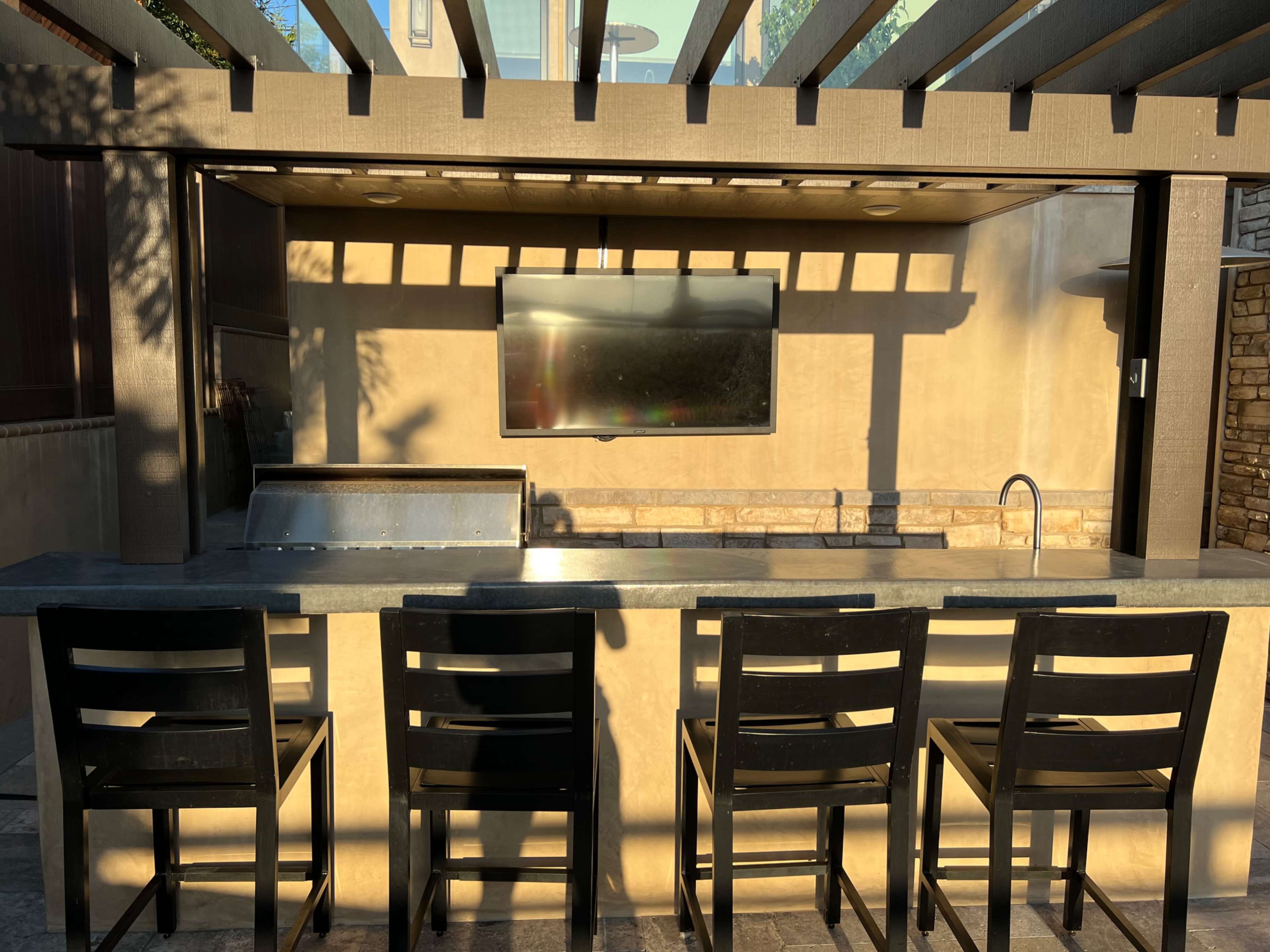 An outdoor bar area features a countertop with four black chairs, a grill, a sink, and a mounted television under a pergola.