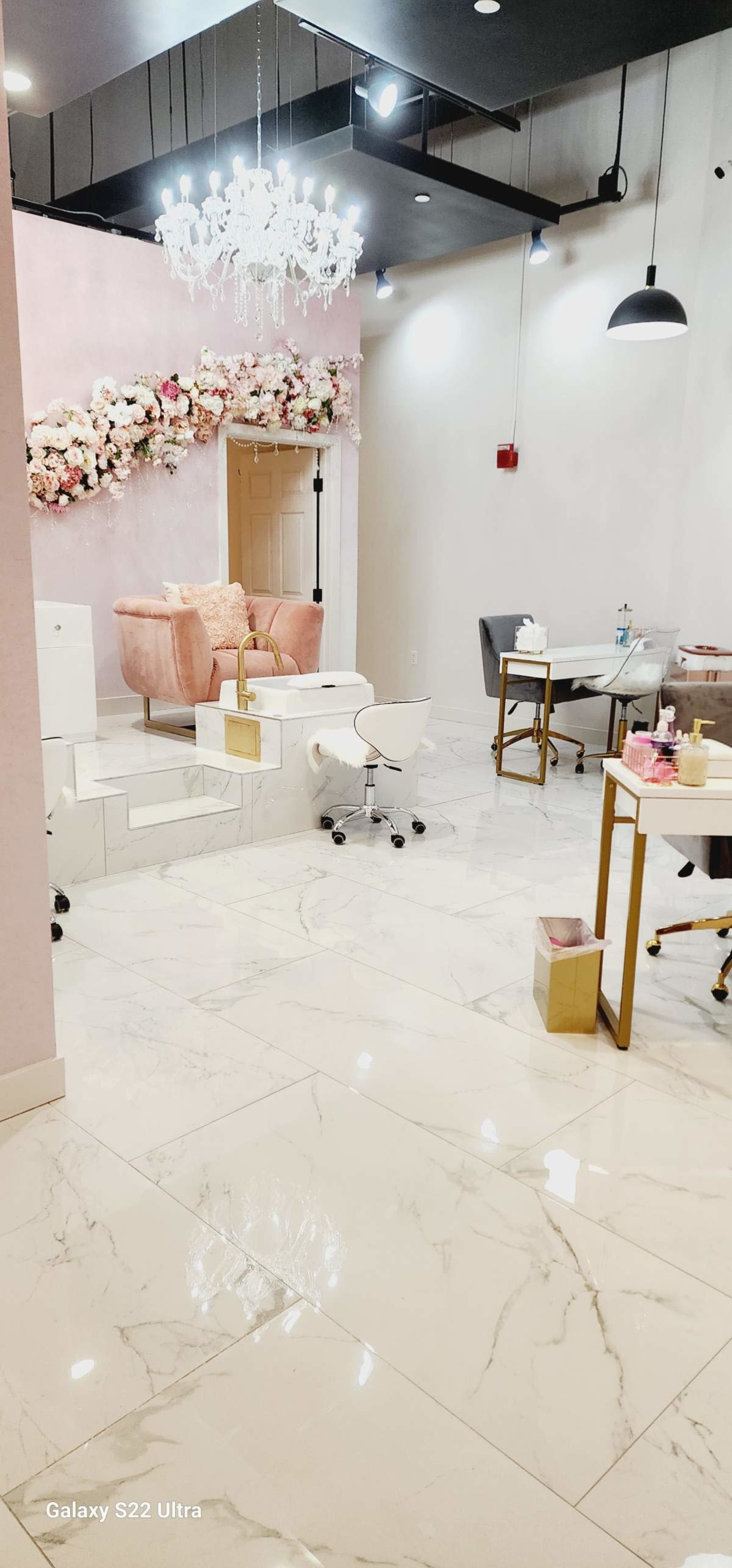 A modern salon interior with marble flooring, pink accent walls, a chandelier, and a floral arrangement above a seating area.