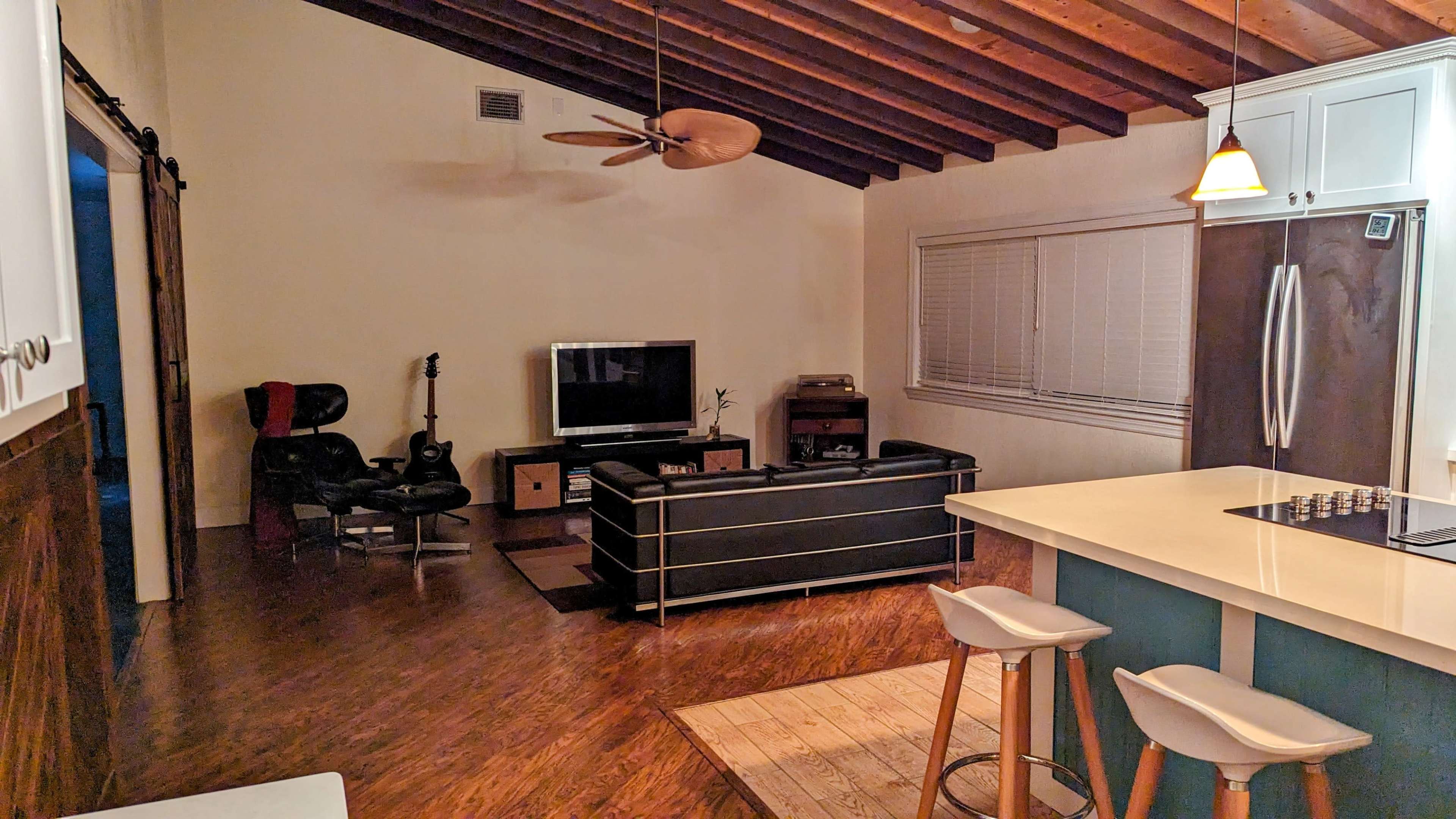 A living space features a black leather couch, a television, a ceiling fan, and a kitchen island with bar stools under wooden beams.