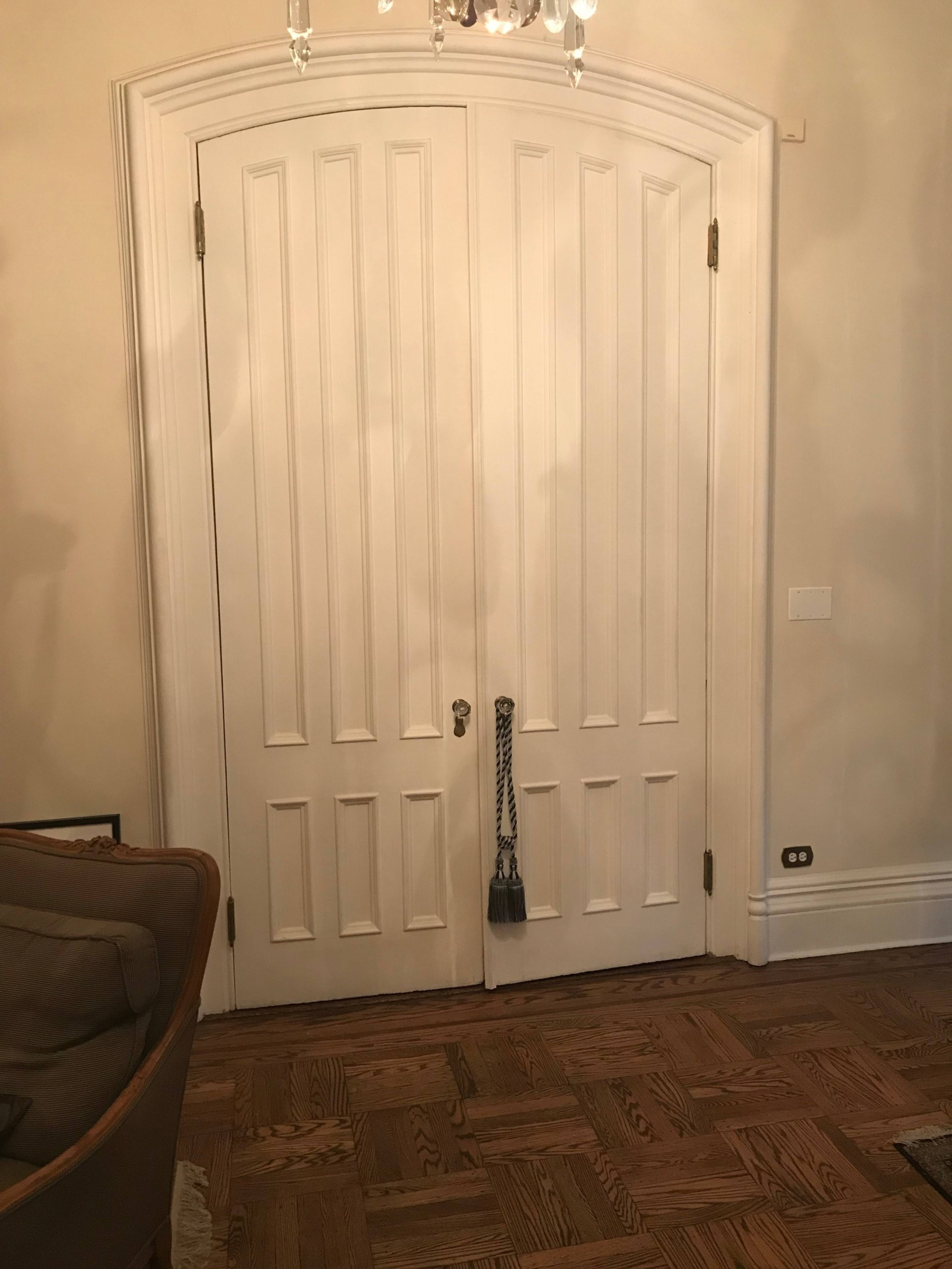 A pair of arched double doors with vertical panels, positioned against a light-colored wall, in a room featuring hardwood flooring.