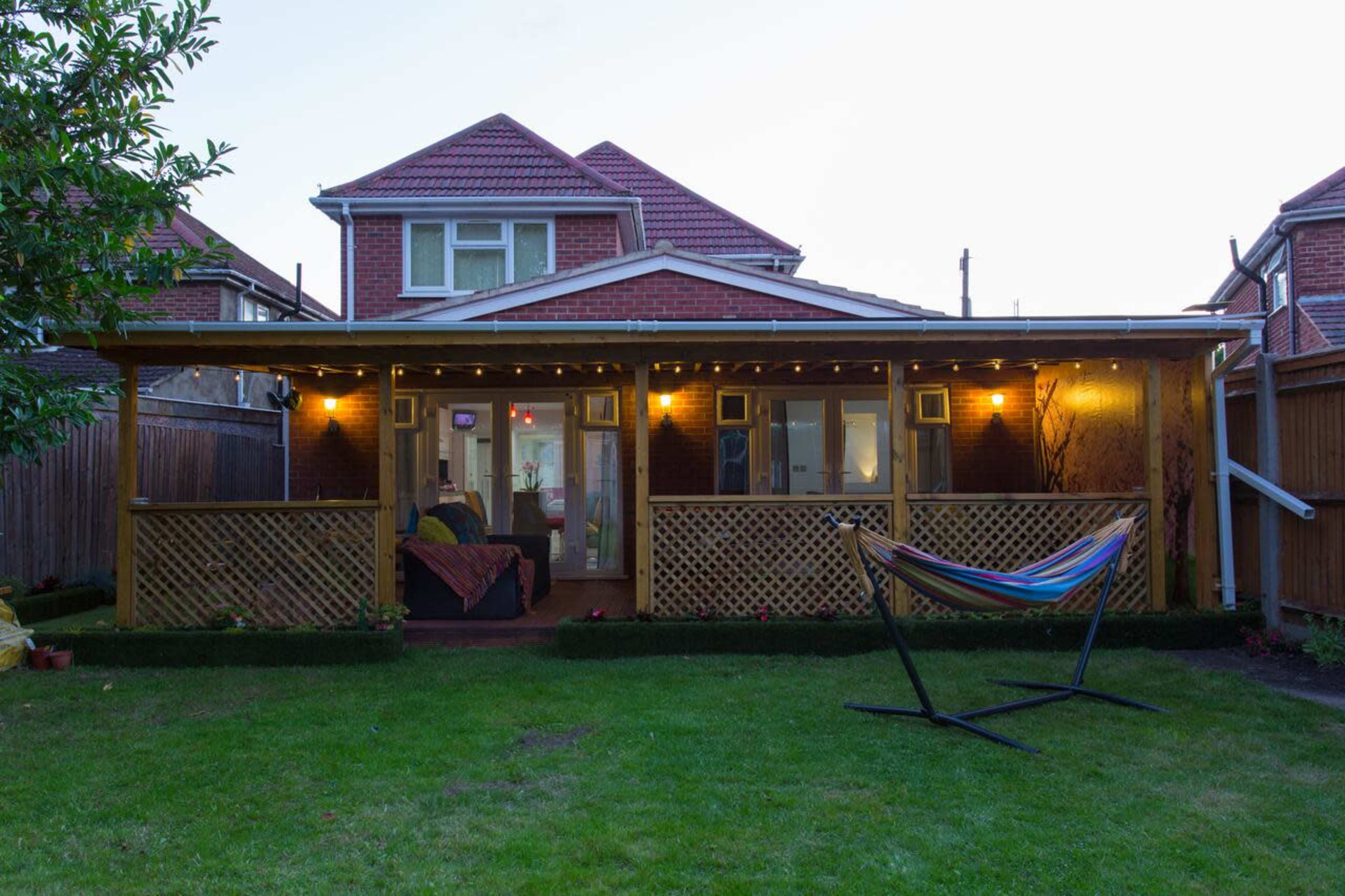 A backyard with a wooden porch illuminated by lights, a hammock stand, and a house in the background.