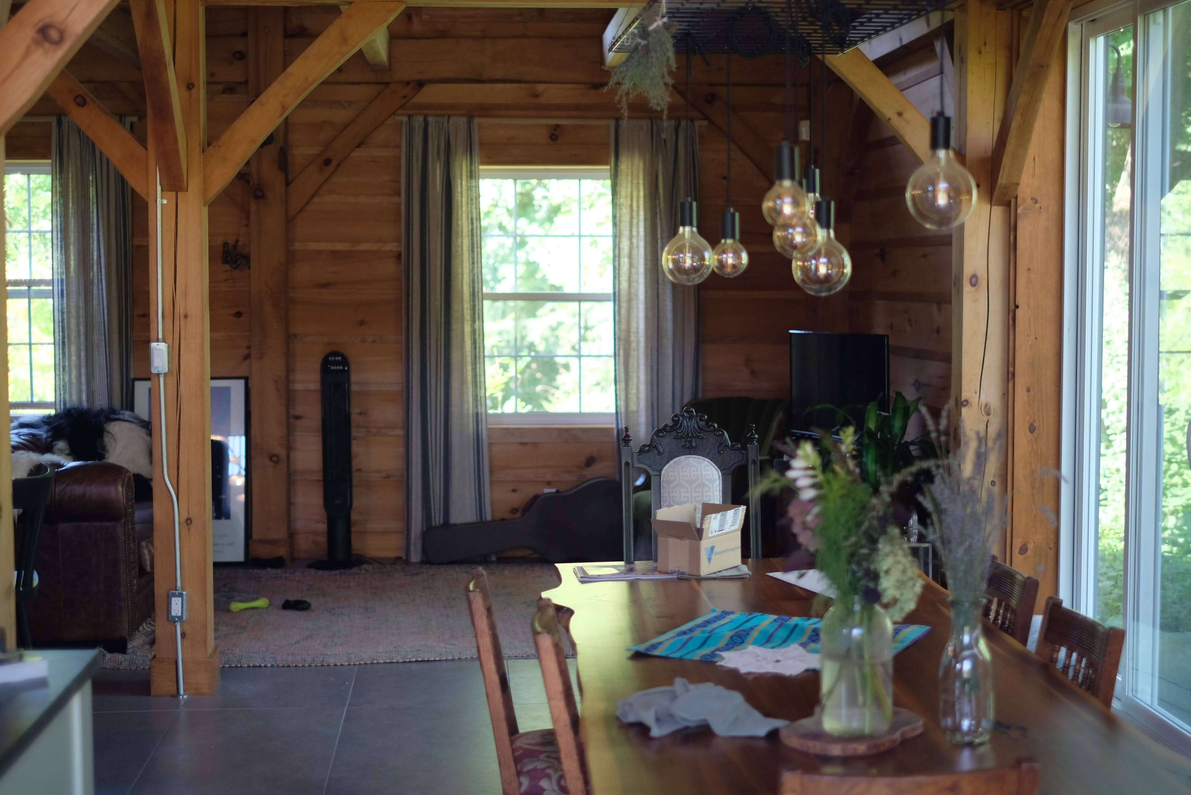 A wooden interior of a living space featuring a dining table with flowers, illuminated by pendant lights, and a view of a cozy lounge area with a sofa.