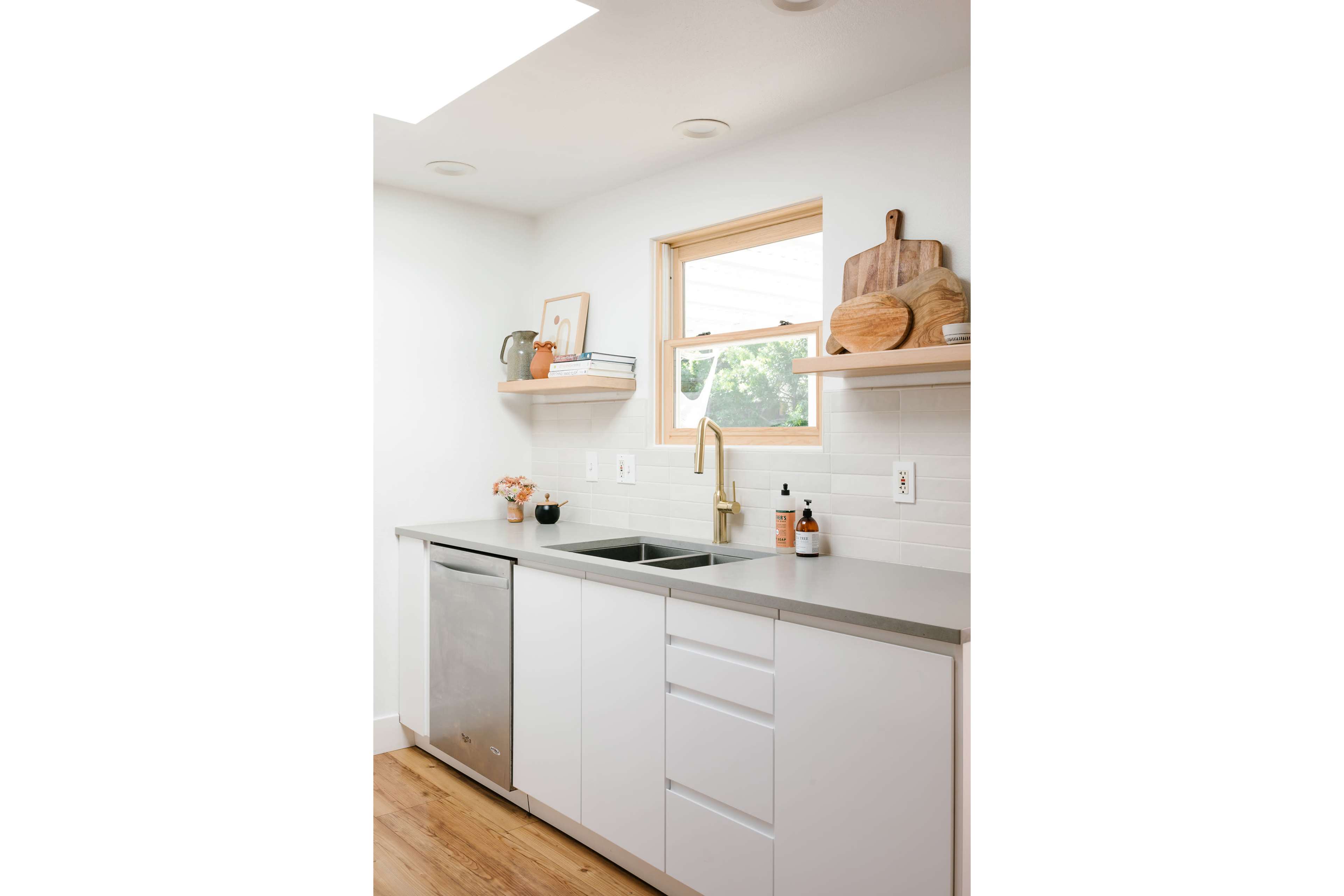 A modern kitchen features a stainless steel dishwasher, a double sink with a gold faucet, and shelves displaying decor and kitchenware.