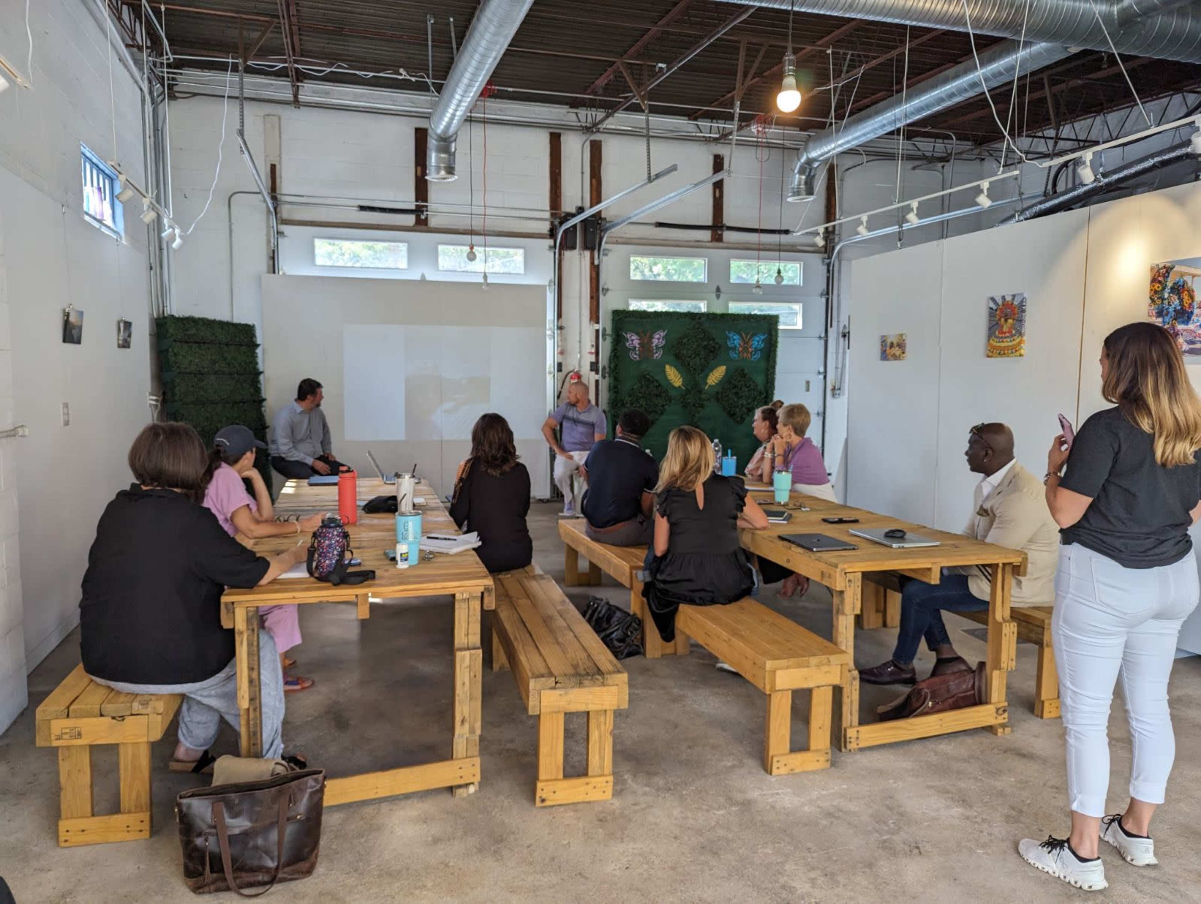 A group of people sits around wooden tables in a bright, industrial-style workspace during a meeting or presentation.