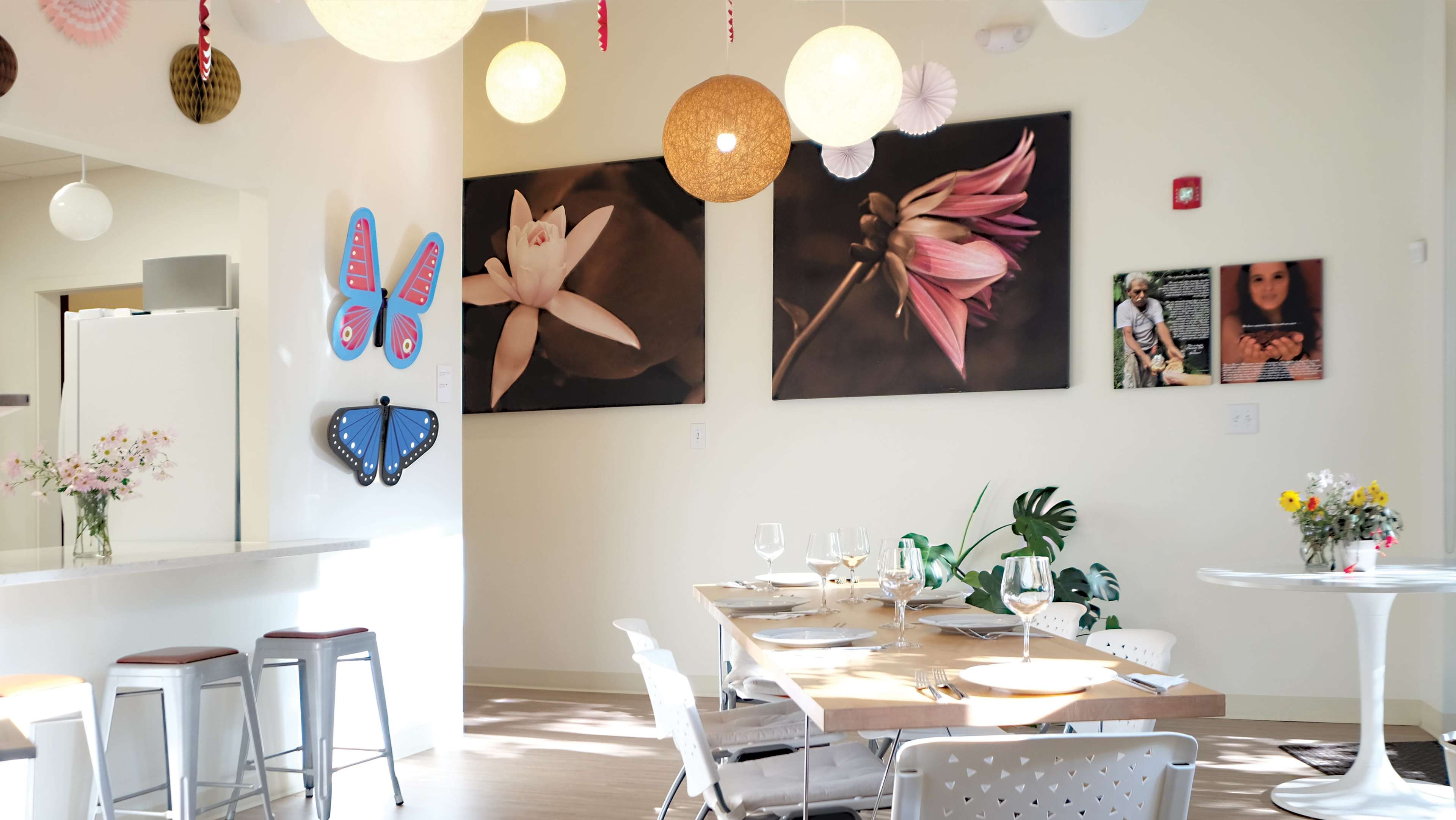The scene shows a brightly lit dining area featuring a table set for a meal, adorned with flower arrangements and surrounded by colorful wall art and decorative lighting.