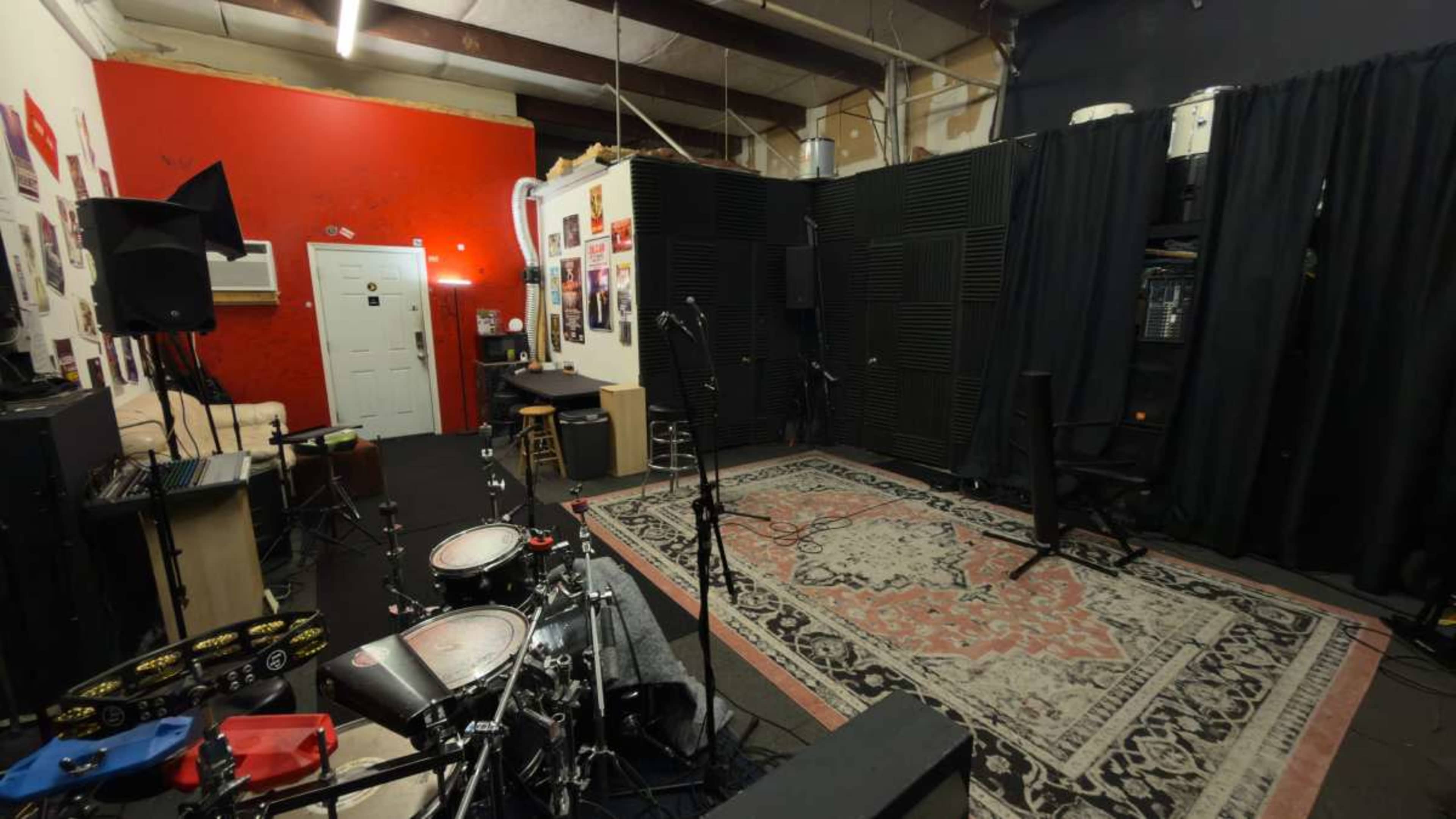 The image shows a music rehearsal studio with a drum set in the foreground, soundproofed walls, and various equipment arranged around a carpeted area.
