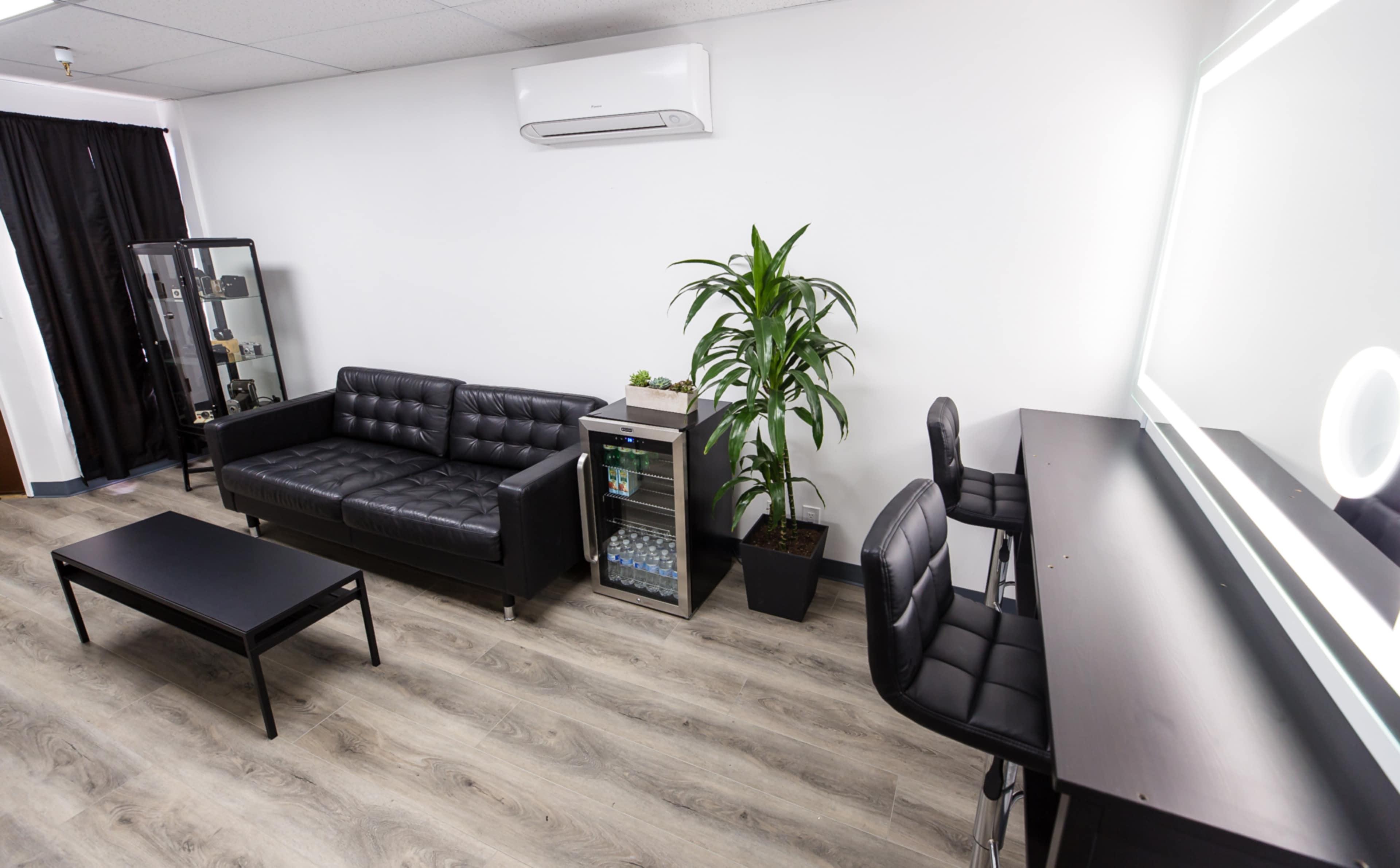 A modern, minimalist waiting area with a black leather couch, a glass display cabinet, a potted plant, a beverage cooler, and a mirrored vanity table with black chairs.