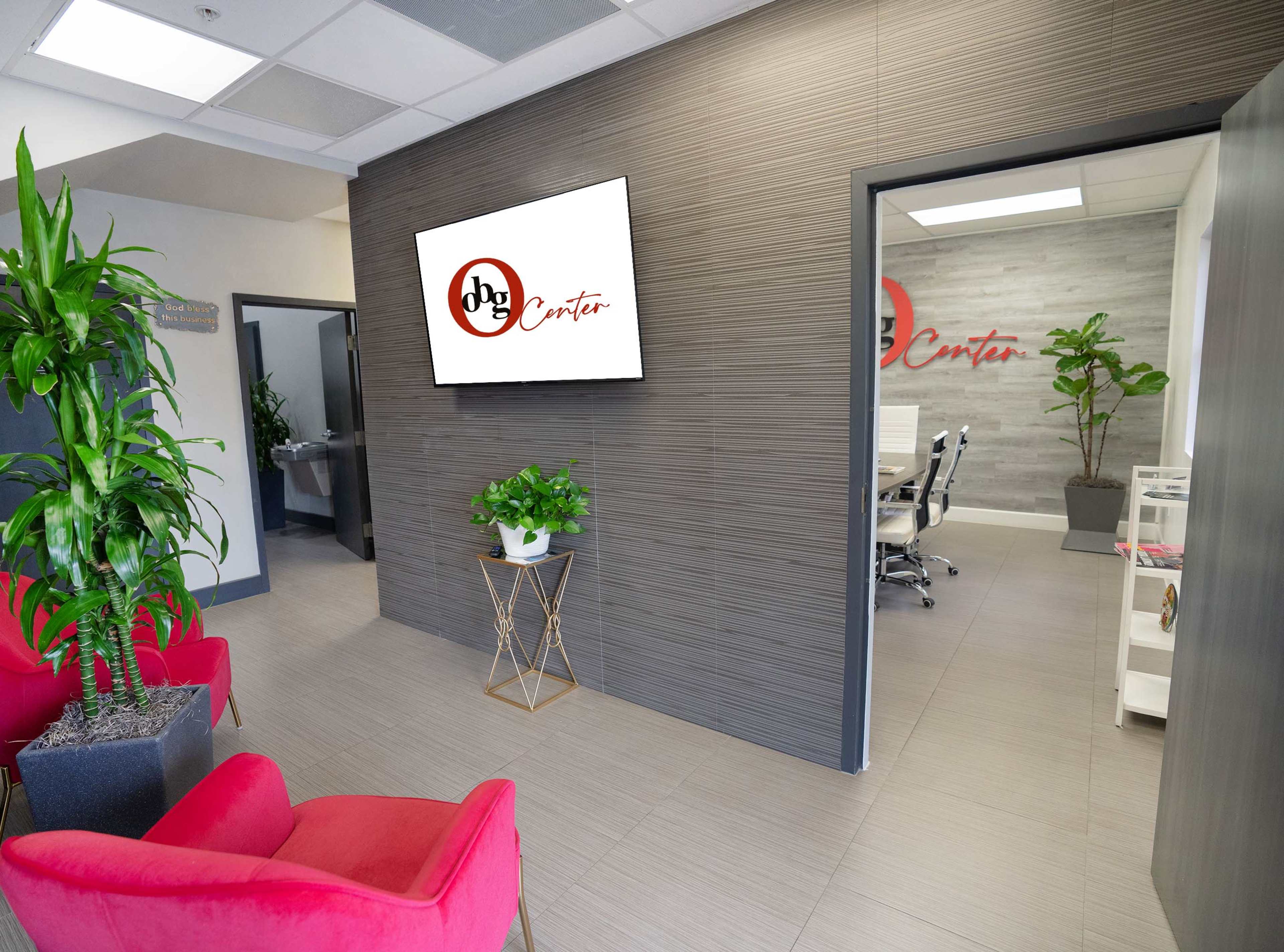 The image shows a modern reception area with a potted plant, a red chair, and a wall-mounted television displaying the logo of a center, leading to a conference room.