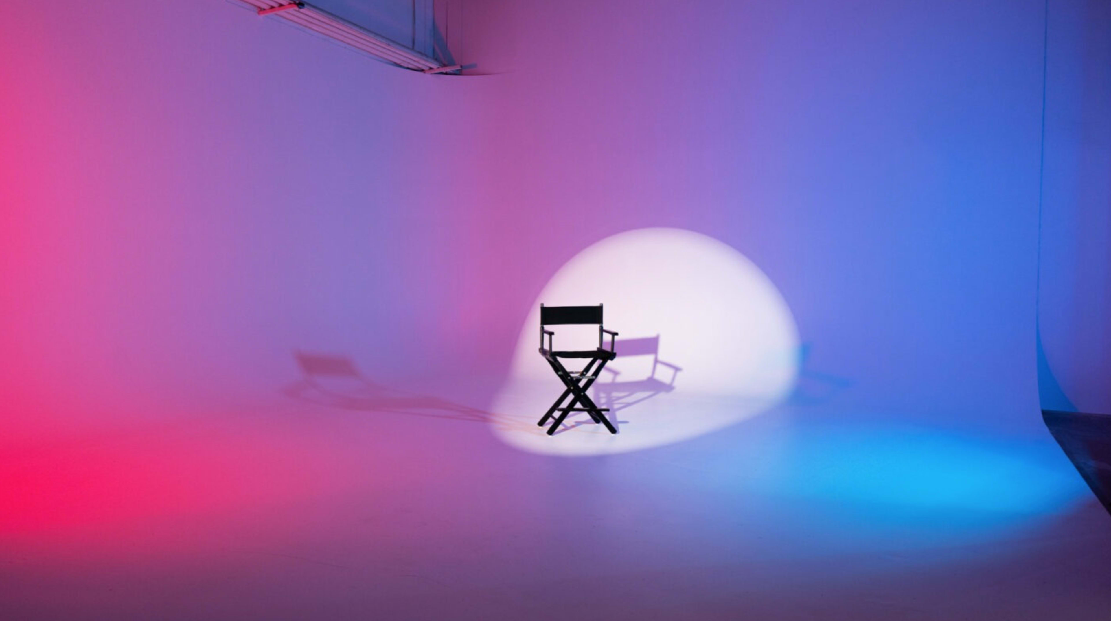 A black director's chair sits in the center of an empty studio with colorful blue and red lighting casting shadows.