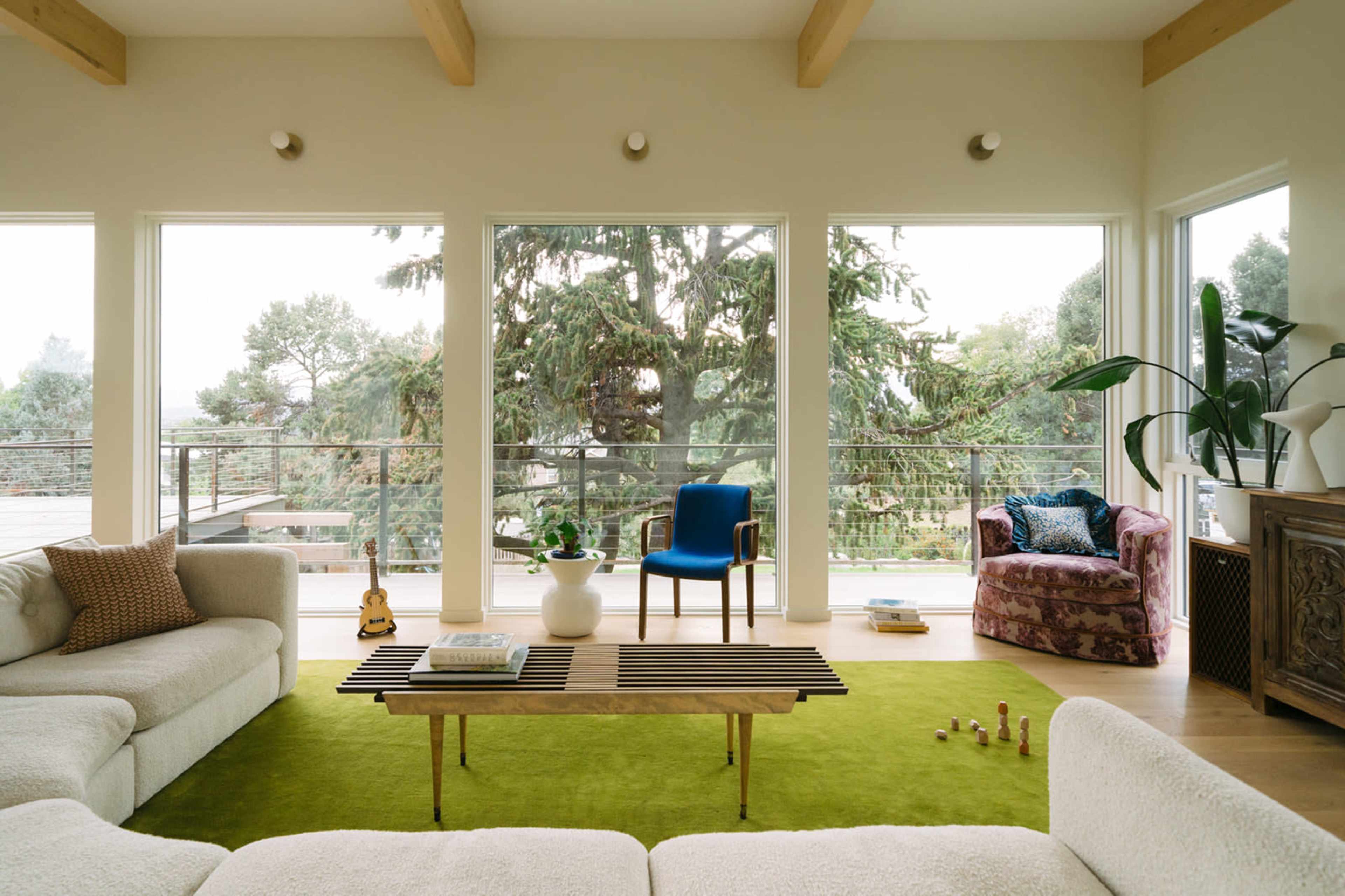 The spacious living room features large windows overlooking greenery, a green rug, and modern furniture arranged for comfort.