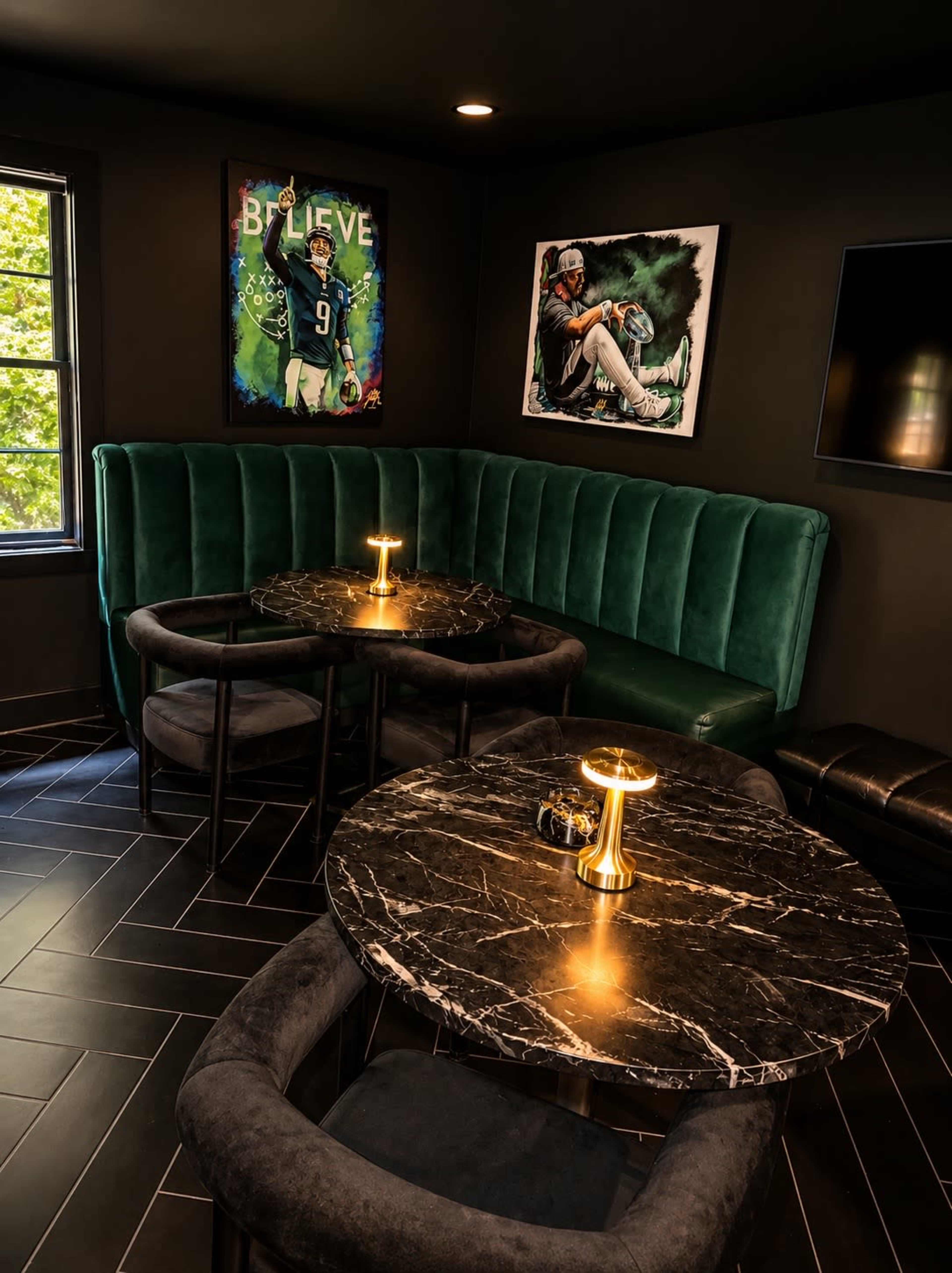 The image shows a modern lounge area with dark walls, a green velvet couch, two round marble tables, and artwork hanging on the walls.