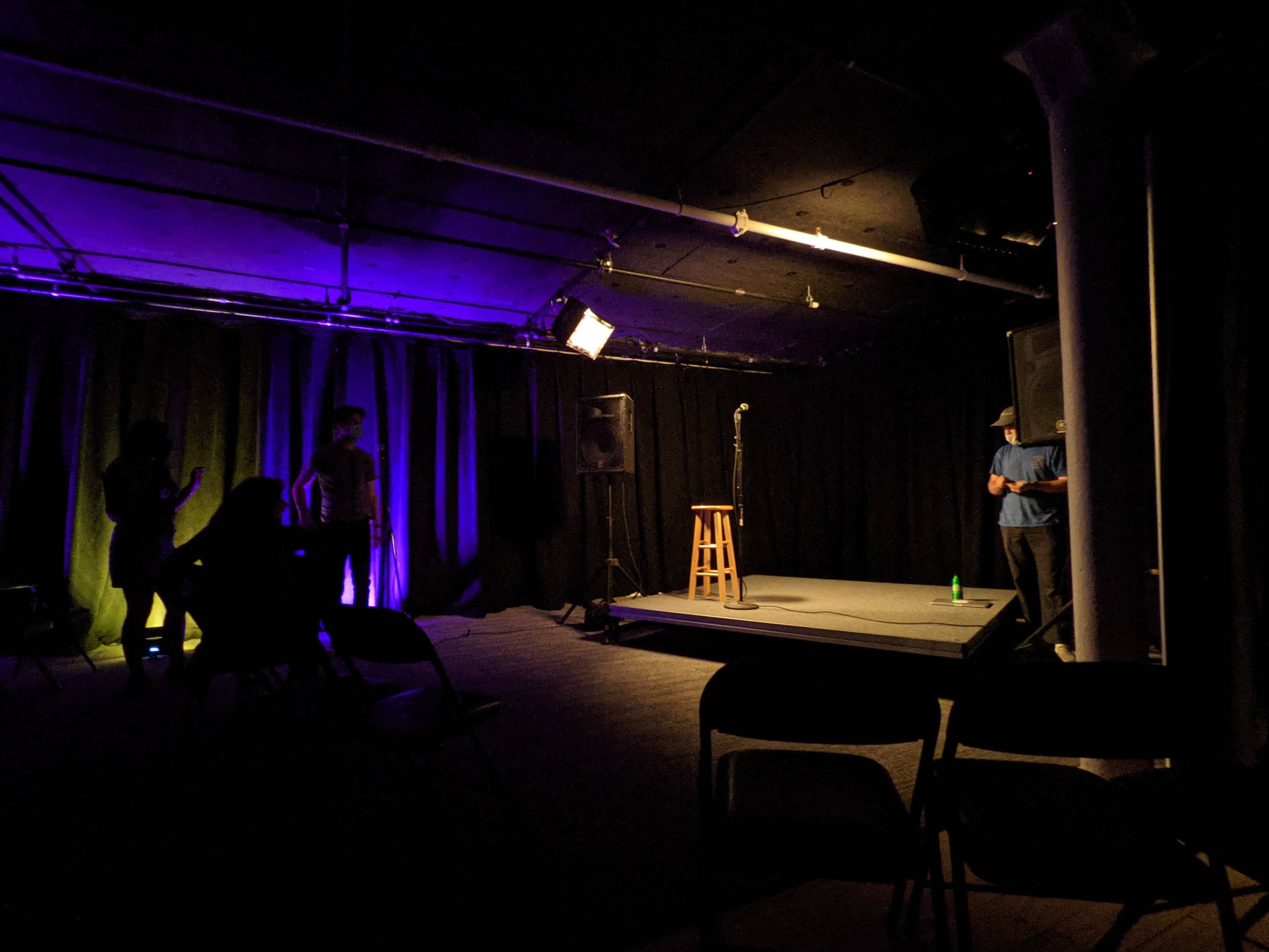 A dimly lit stage with a microphone on a stool, surrounded by empty chairs and silhouettes of people in the background.