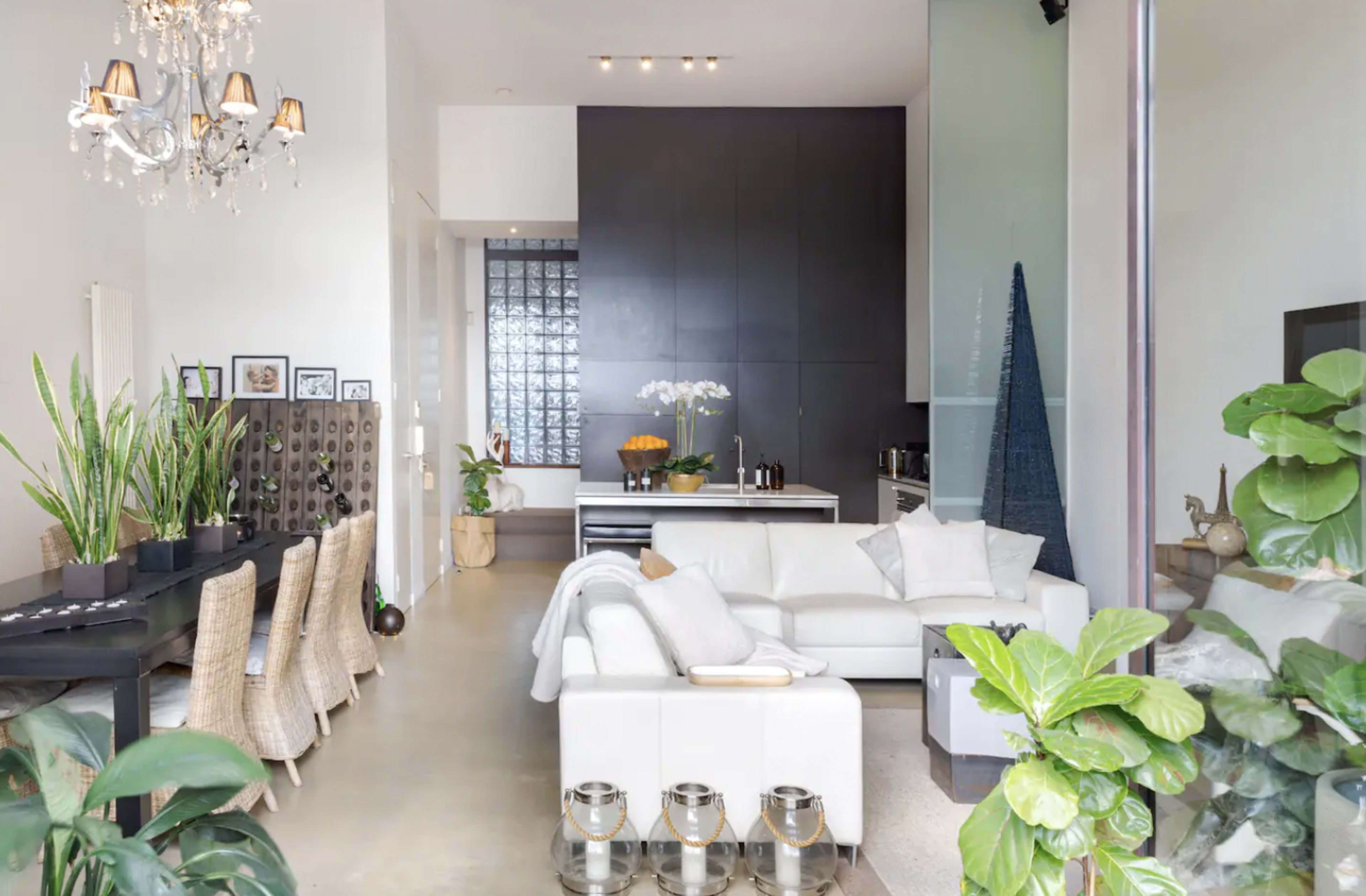 The image shows a modern living space featuring a white sectional sofa, a dining area with woven chairs, and large plants, set against a backdrop of sleek furniture and a glass block wall.