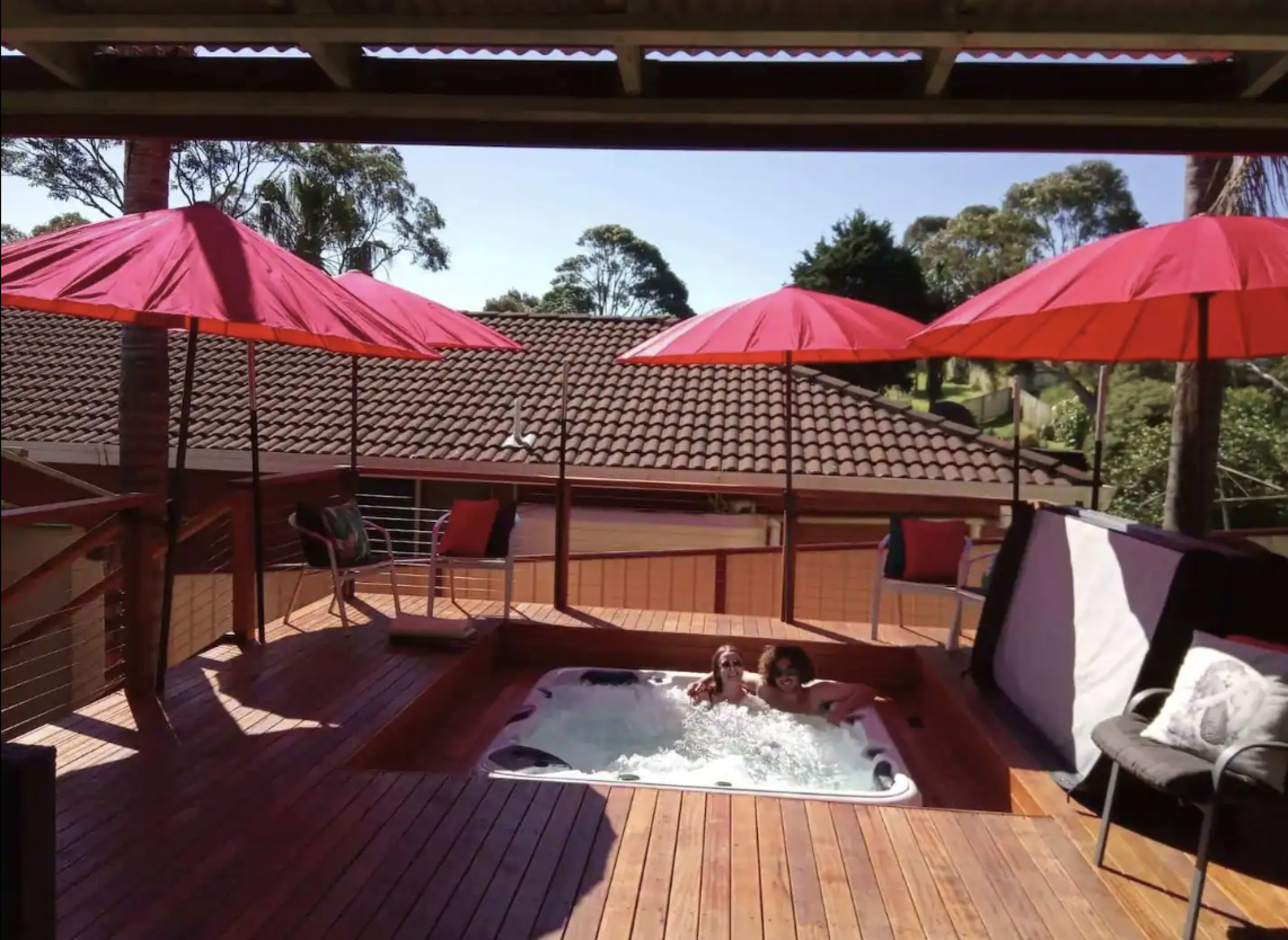 Two people in a hot tub on a wooden deck surrounded by red umbrellas overlooking a residential area.
