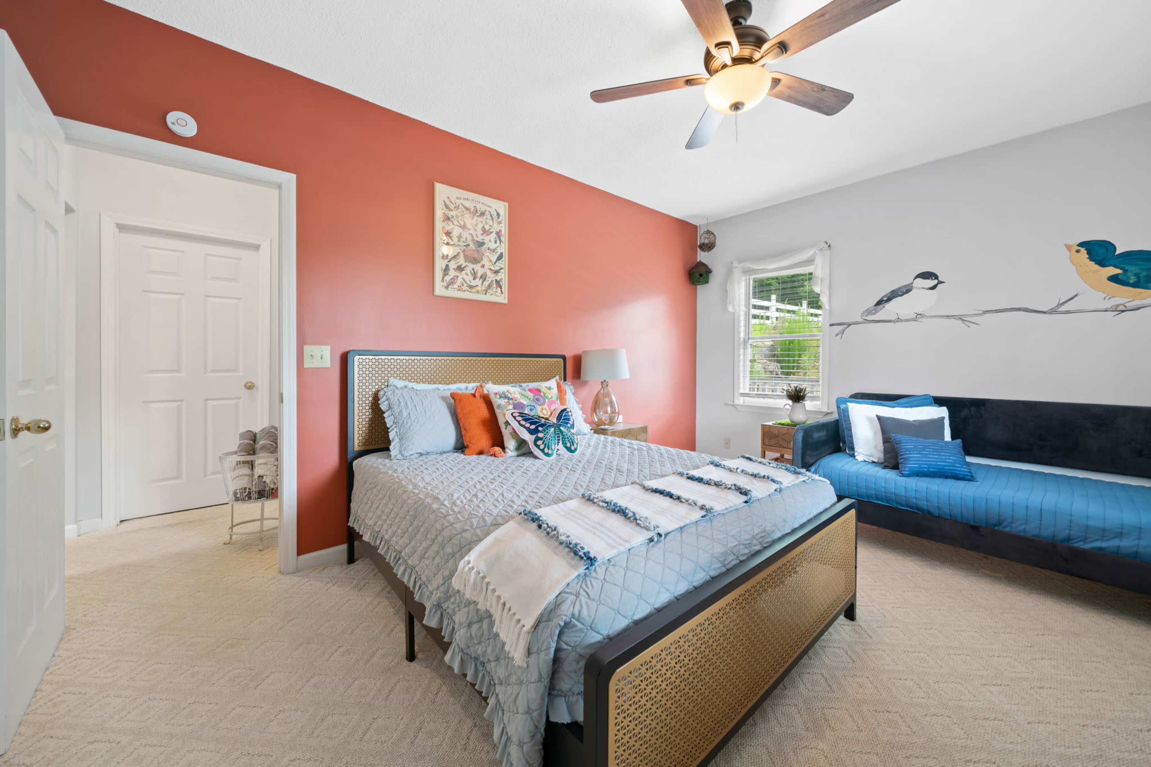 The image shows a bedroom featuring a bed, a couch, and a mural of birds on the wall, with an adjoining door to a bathroom visible.