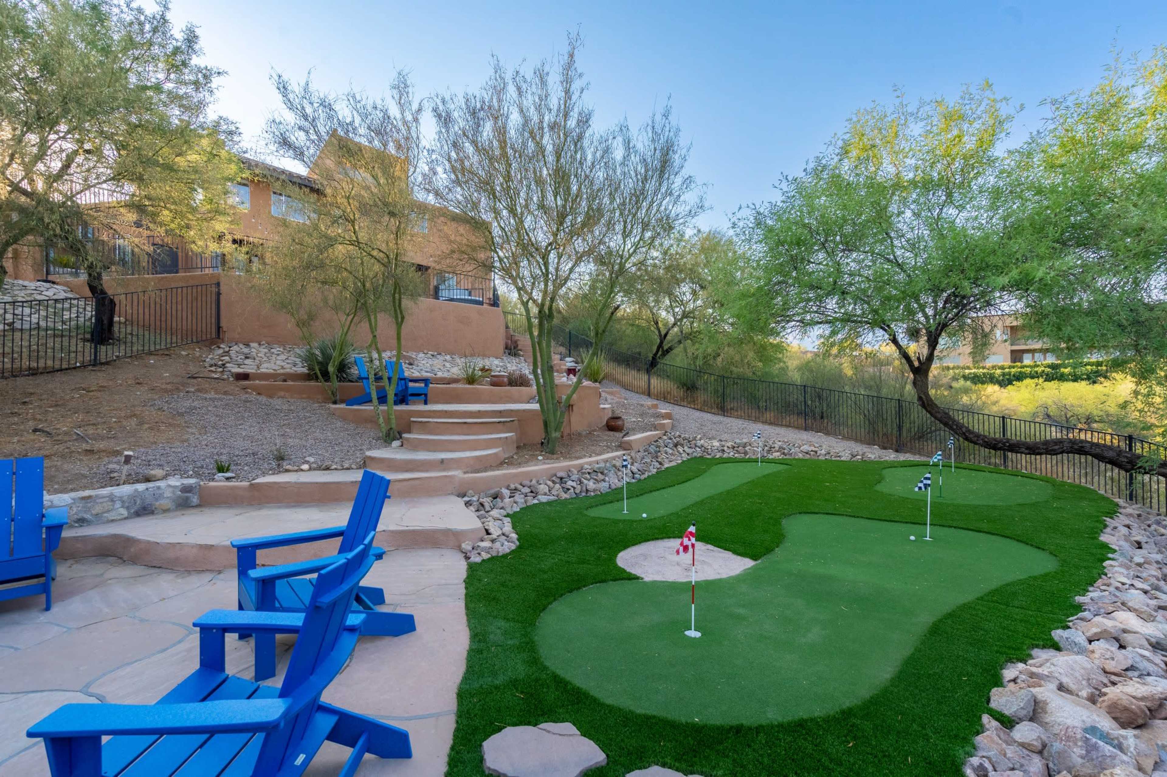 A landscaped backyard featuring a putting green surrounded by shrubs, rocks, and a set of blue outdoor chairs.