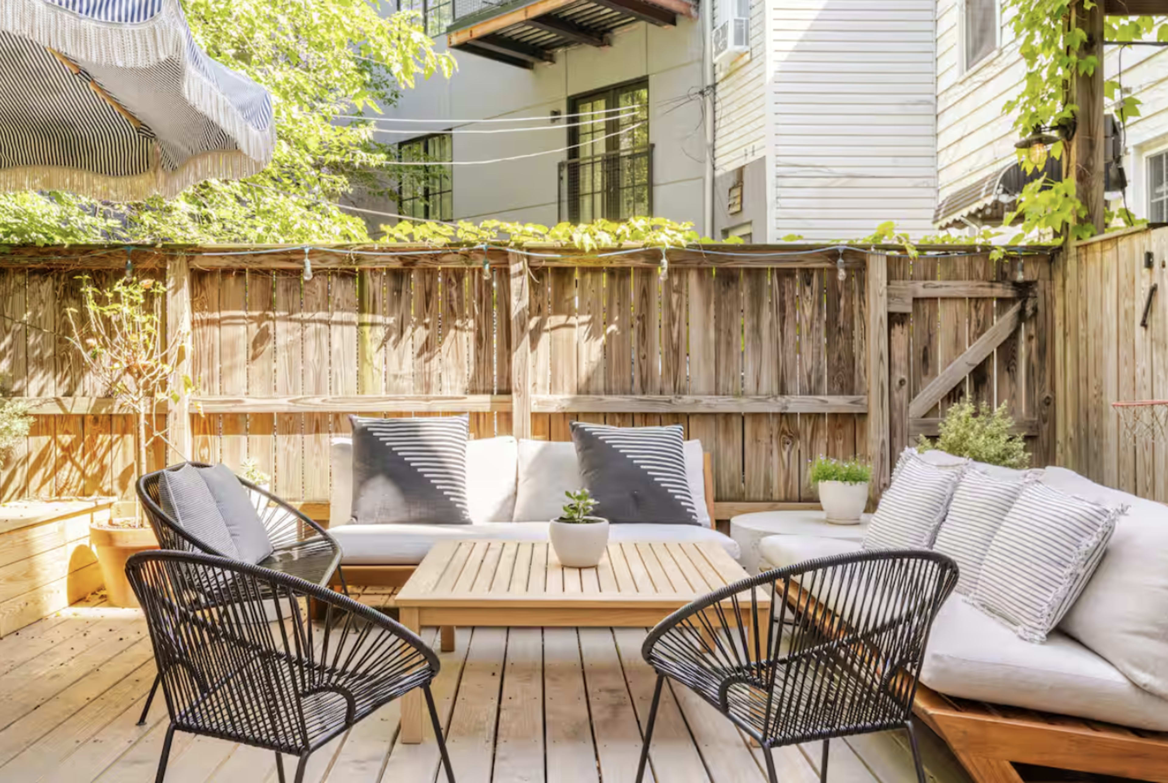 The image shows a small outdoor patio area with wooden decking, featuring a seating arrangement of a wooden table and several black woven chairs, surrounded by a wooden fence and greenery.