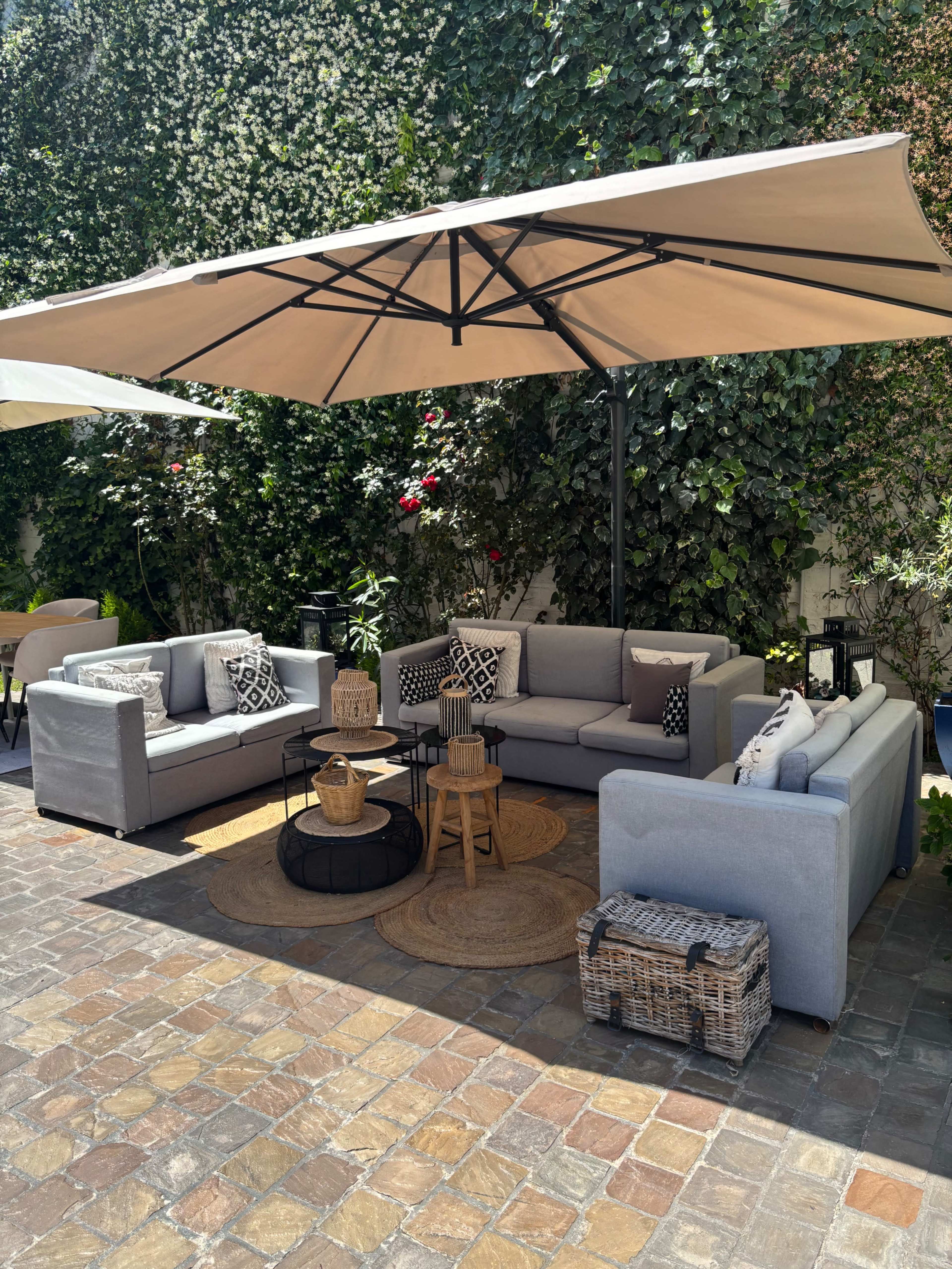 A cozy outdoor seating area features gray sofas arranged around a circular black coffee table, surrounded by lush greenery and shaded by a large umbrella.