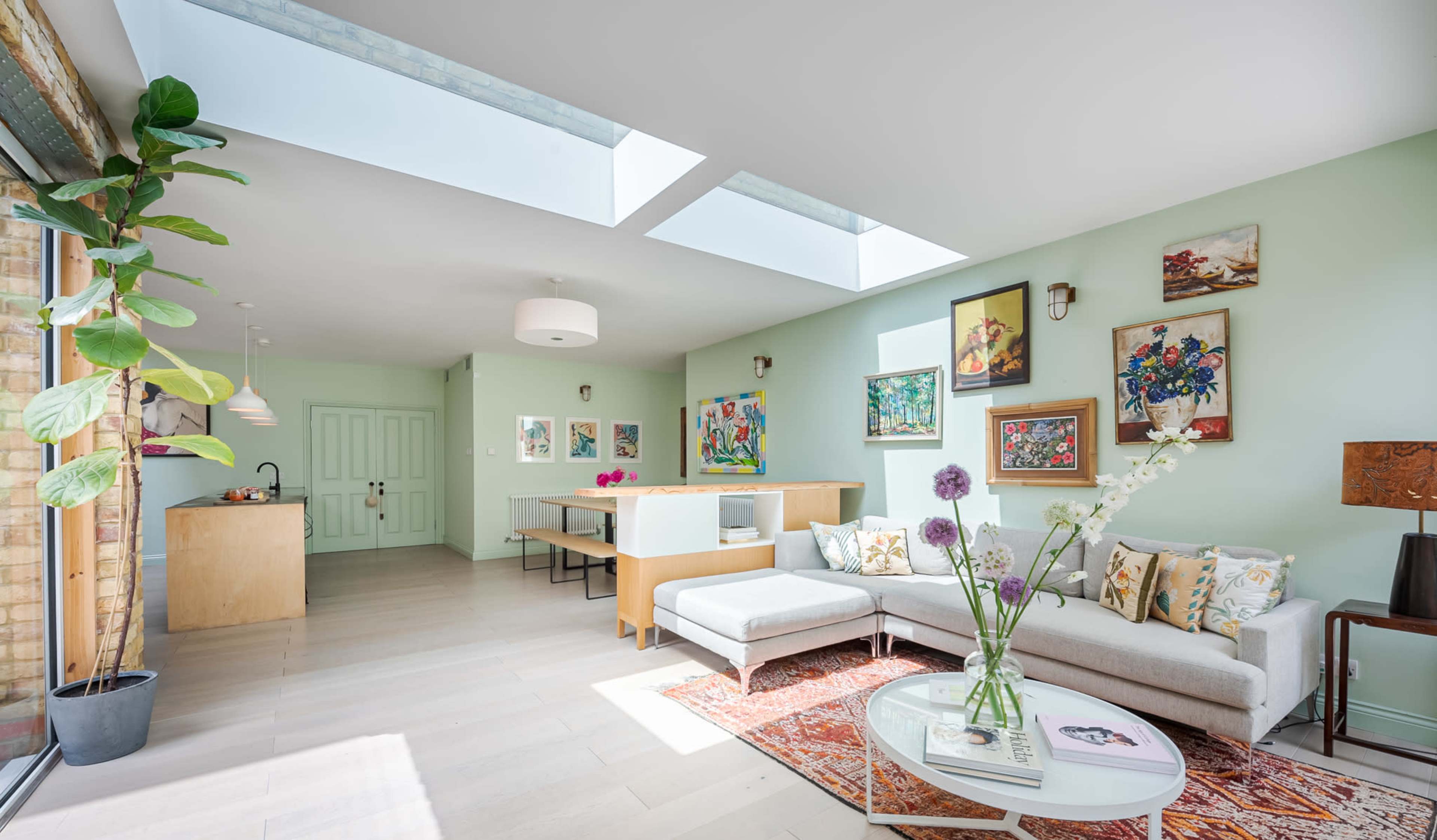 A bright, modern living space featuring a light green color scheme, a sectional sofa, framed artwork on the walls, and large skylights allowing natural light to enter.