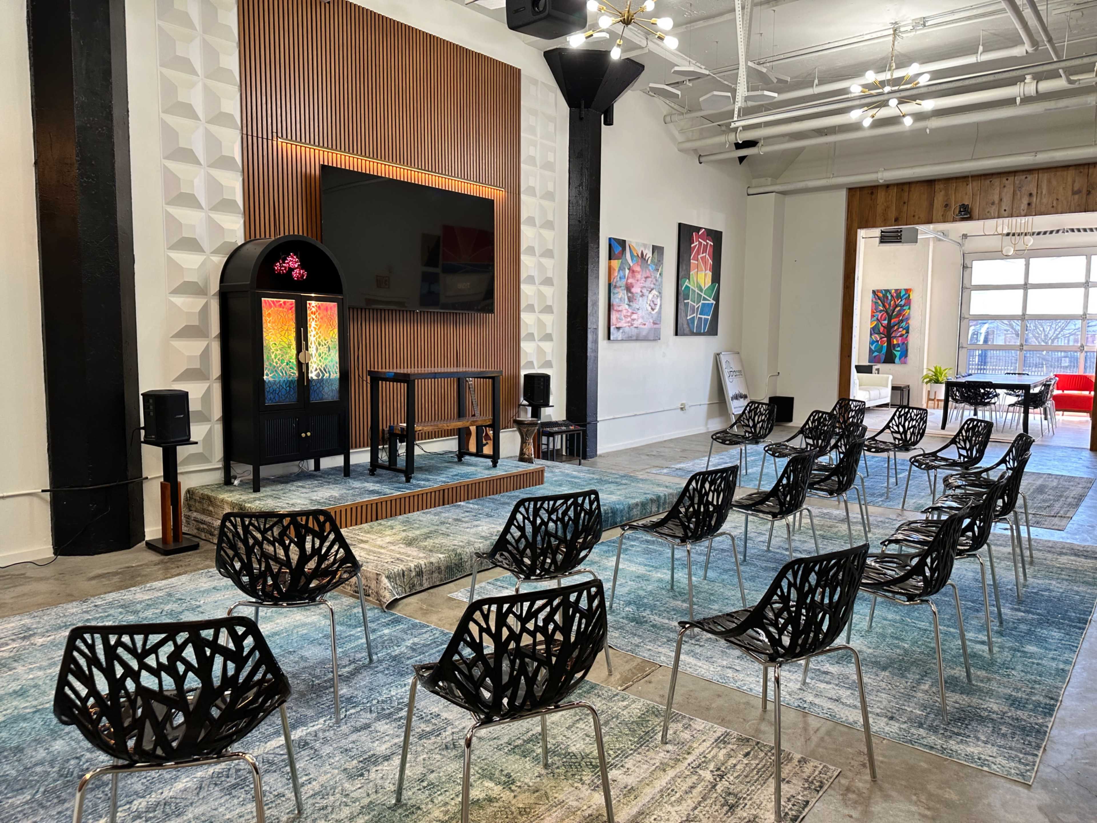 The image shows a spacious room set up for a presentation, featuring rows of black chairs arranged in front of a large screen and colorful artwork on the walls.