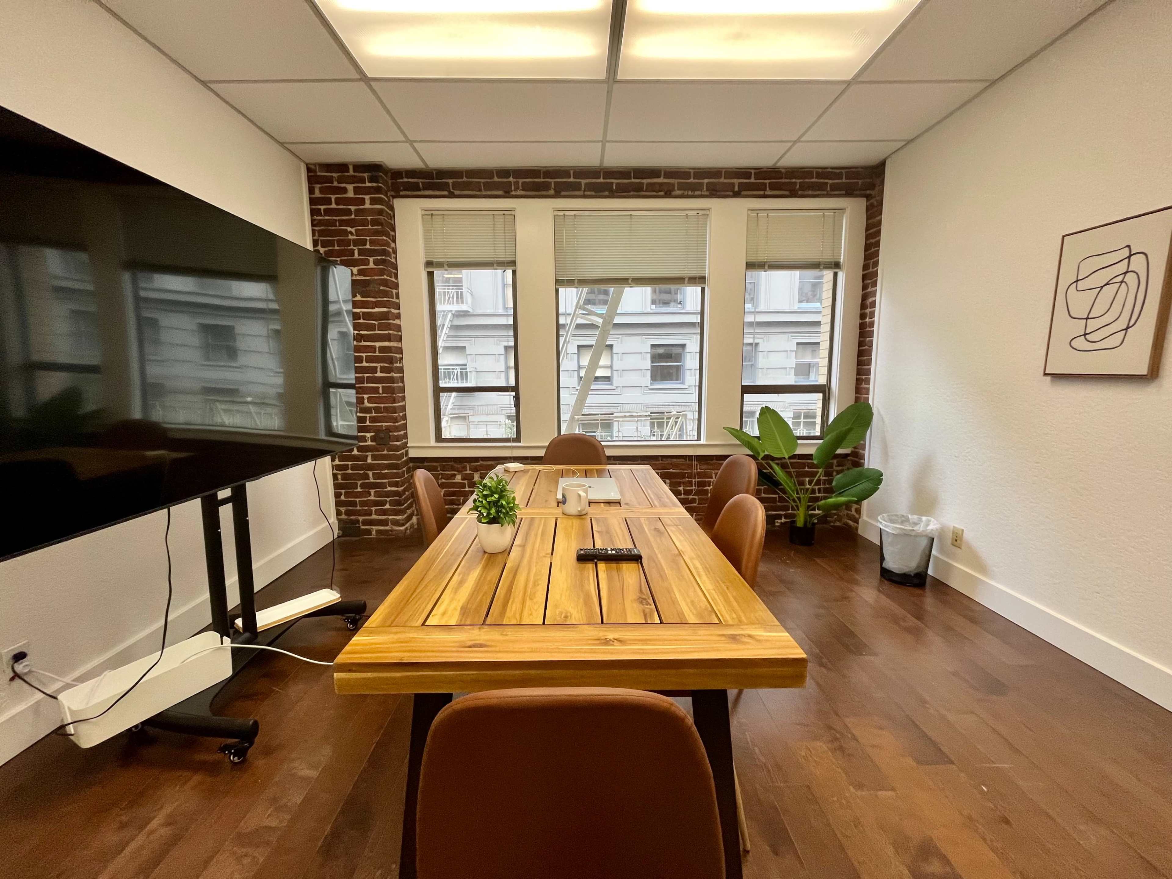 A conference room features a wooden table surrounded by chairs, with a large television screen mounted on one wall and windows looking out onto a building facade.