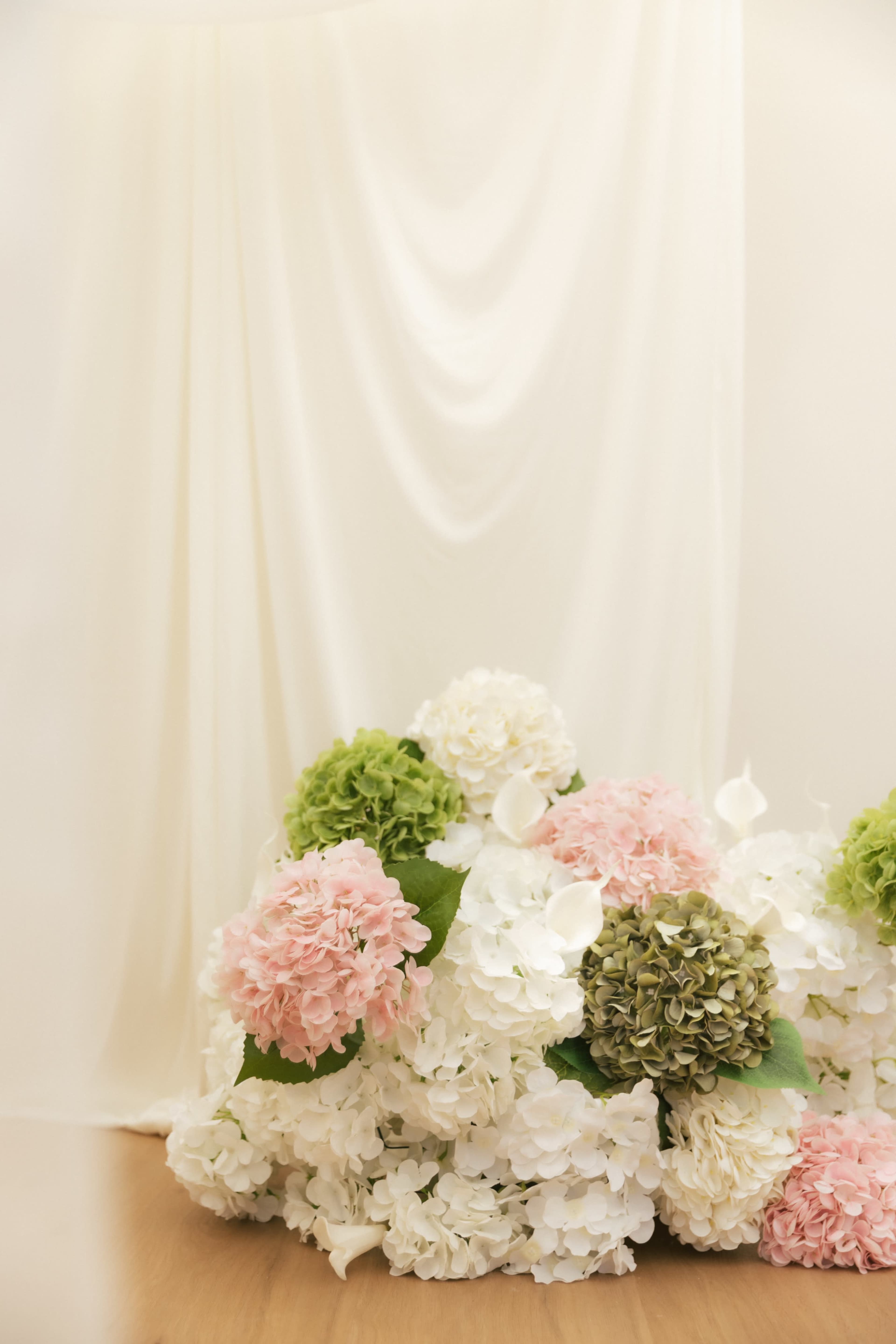 A mass of artificial flowers in shades of pink, white, and green is arranged against a softly draped white backdrop.