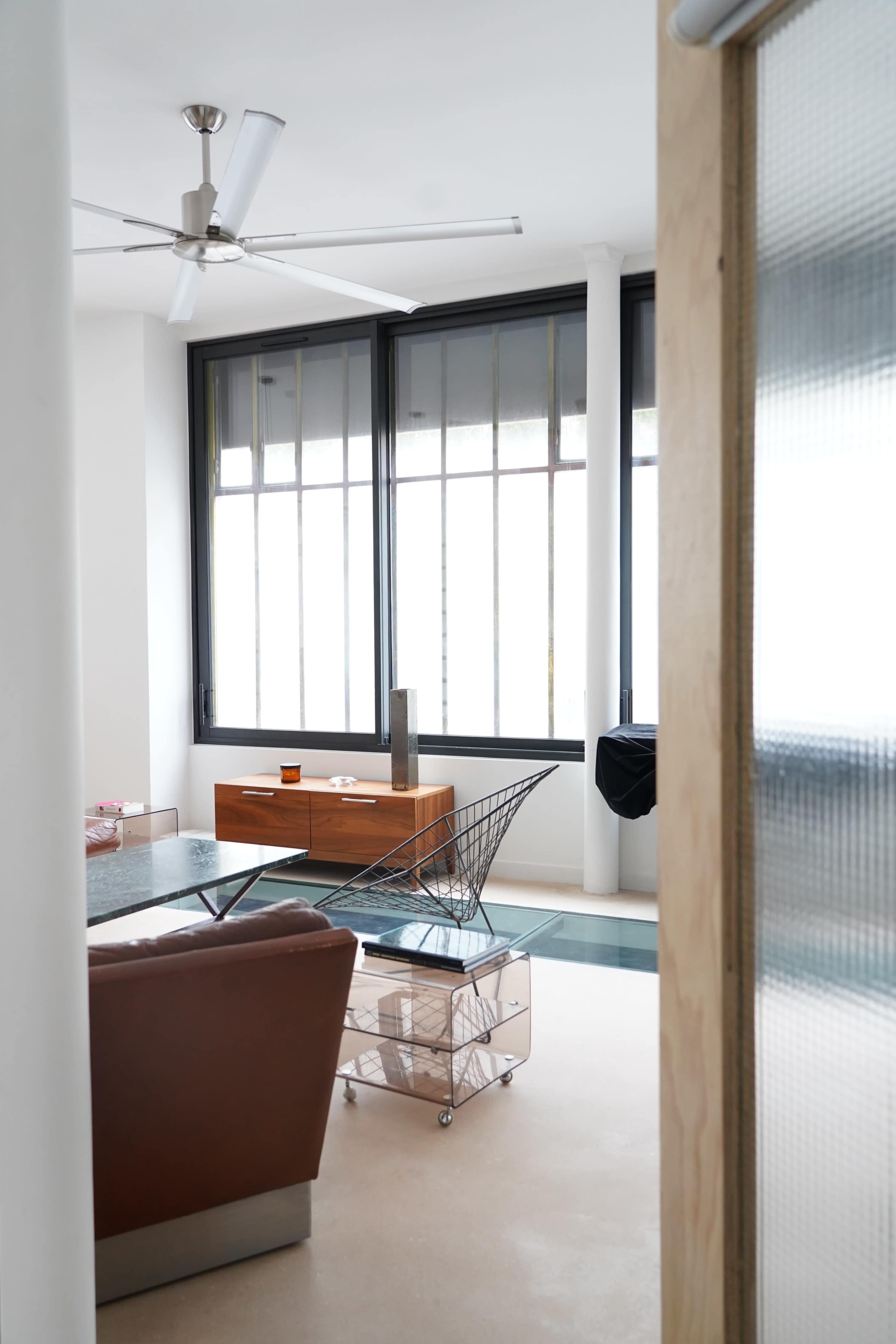 The image shows a modern living room with large windows, a brown leather sofa, and a wire cart beside a glass table.