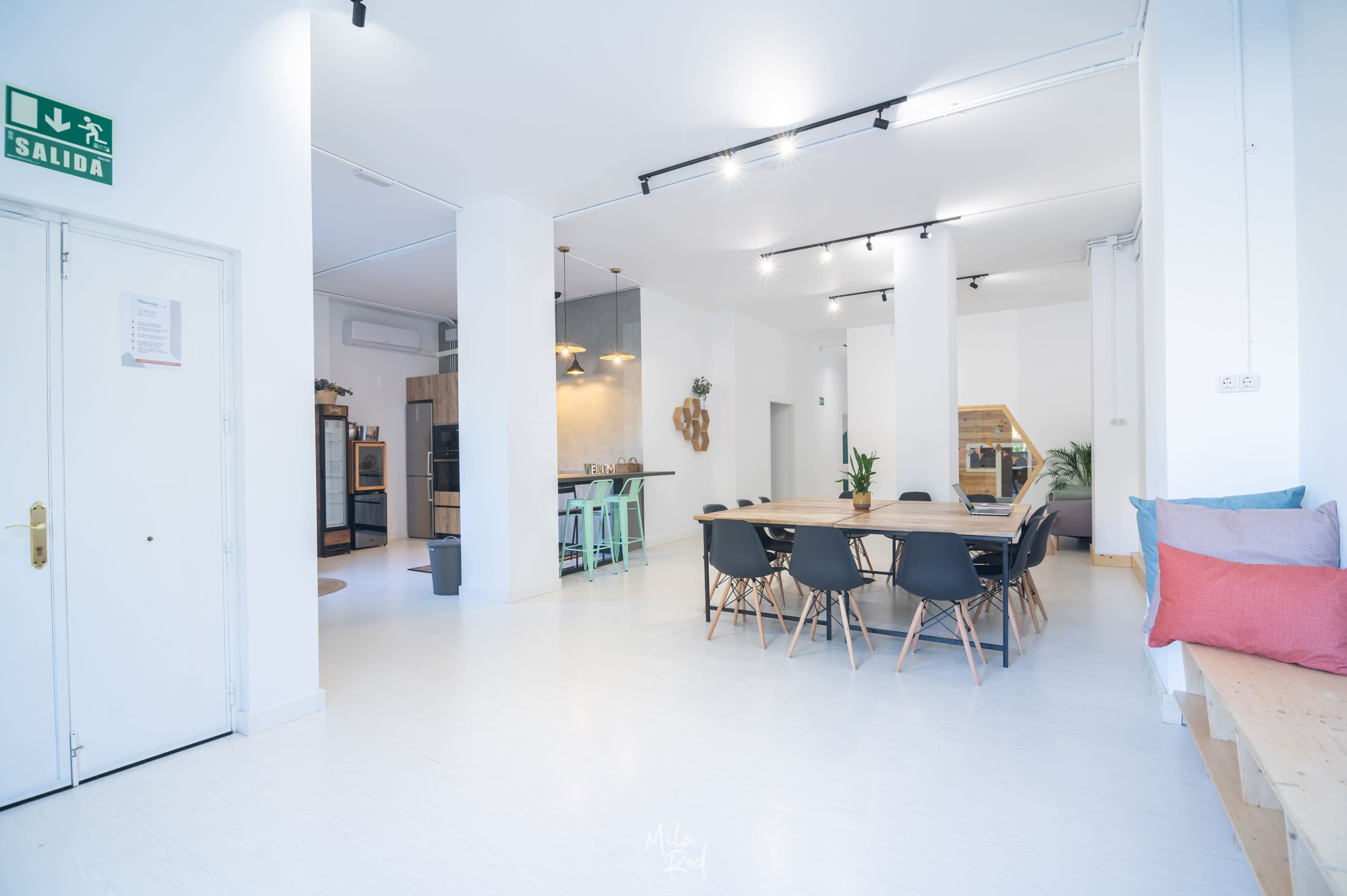A spacious, modern interior with a large dining table surrounded by chairs, a kitchen area in the background, and natural light illuminating the room.