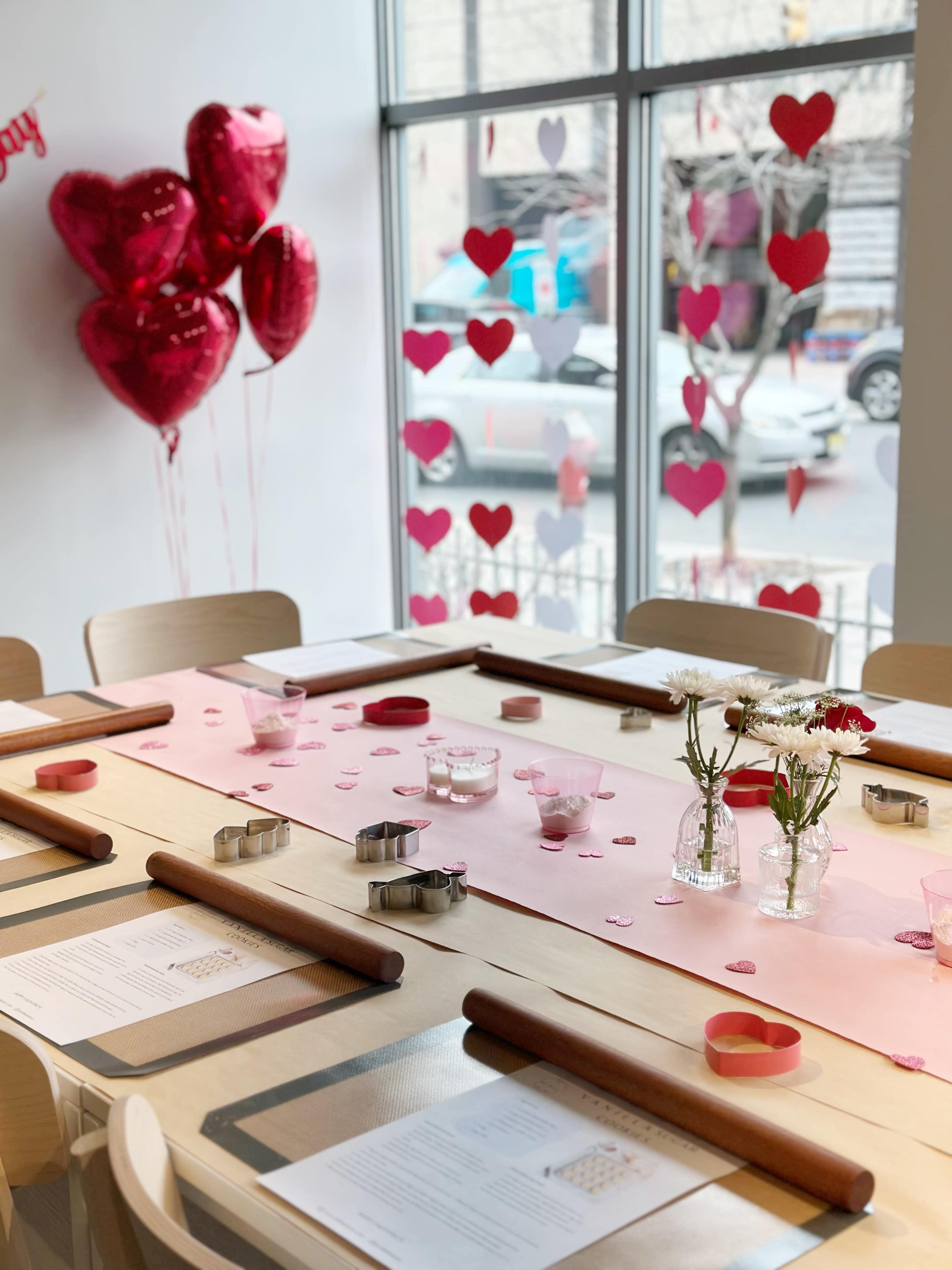 A dining table is decorated with pink tableware and heart-shaped decor, surrounded by heart-shaped balloons and window displays.