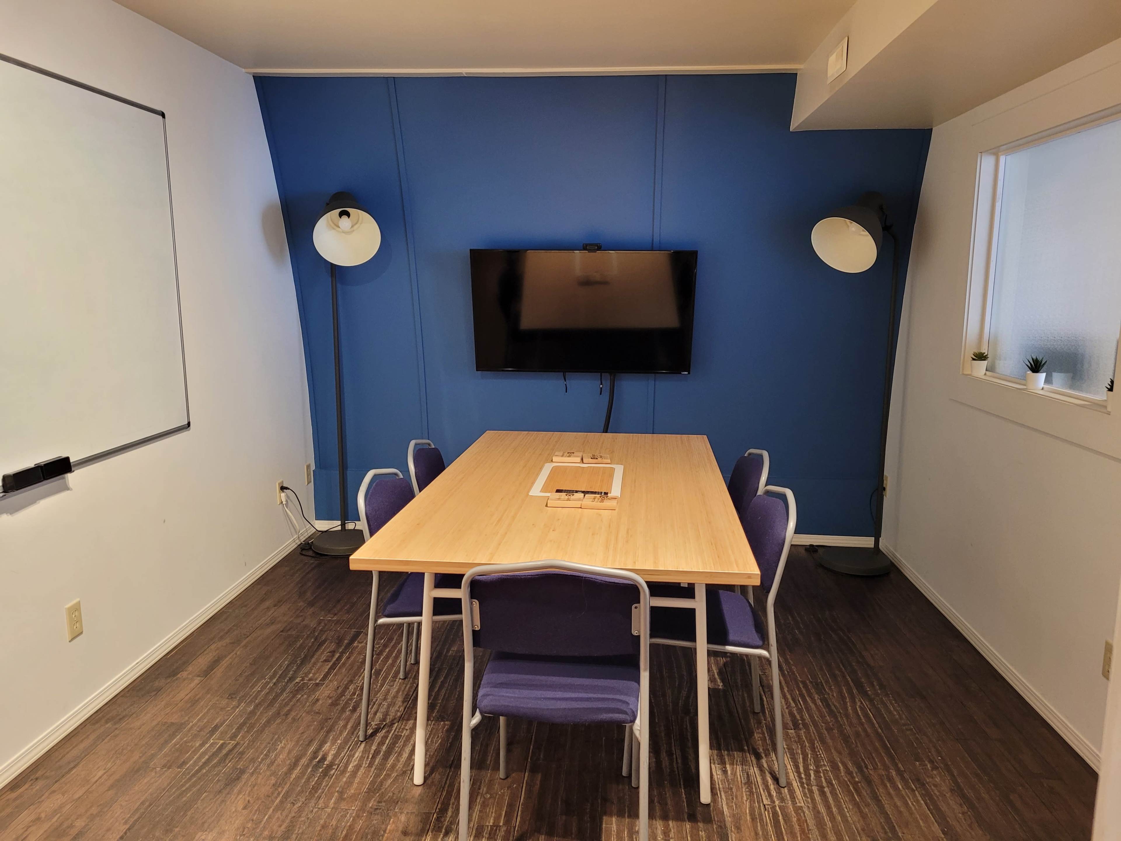 A conference room features a wooden table surrounded by purple chairs, with a blue wall and a television mounted on it.