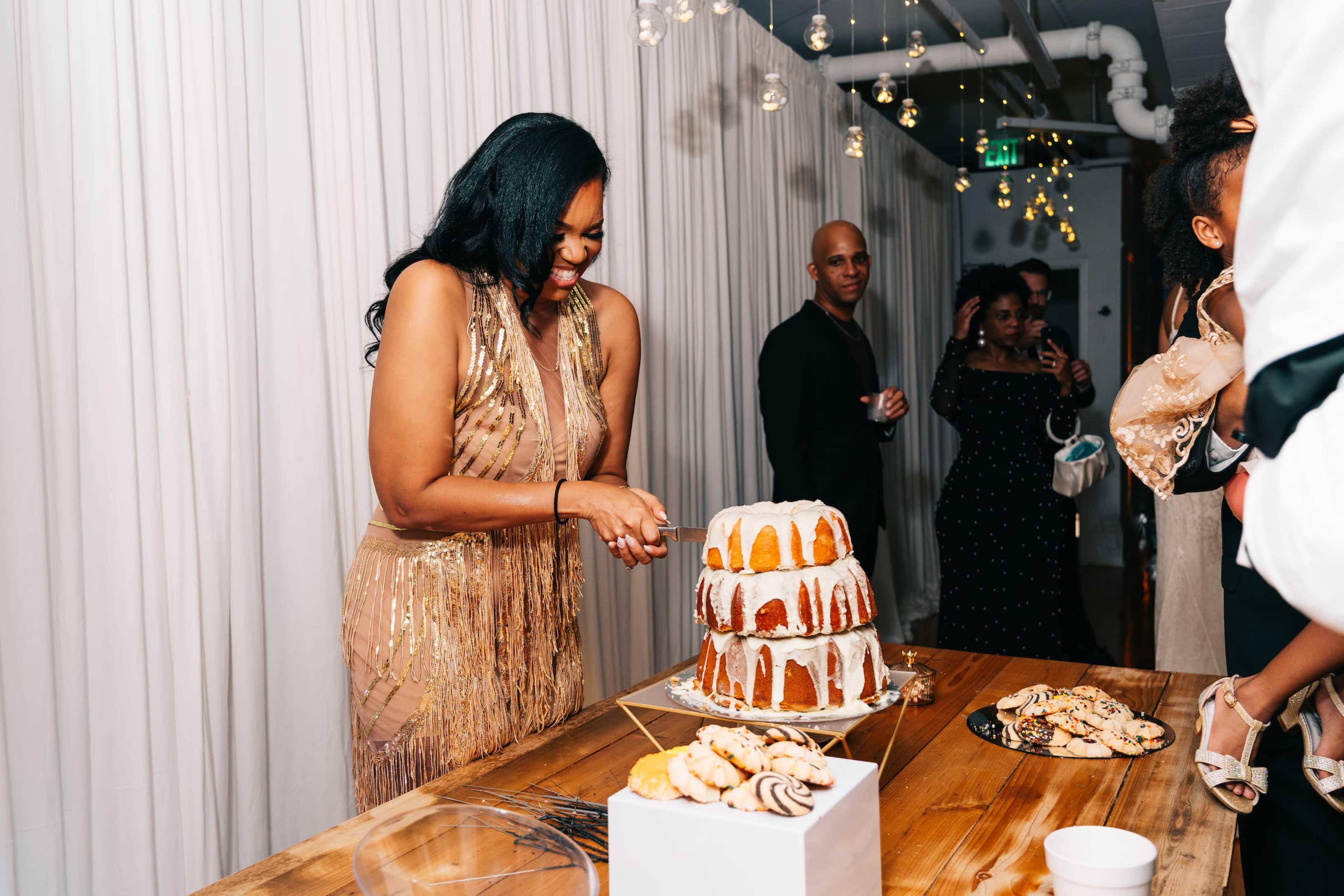 A woman in a sparkling dress cuts a multi-layered cake while guests socialize in a decorated room.