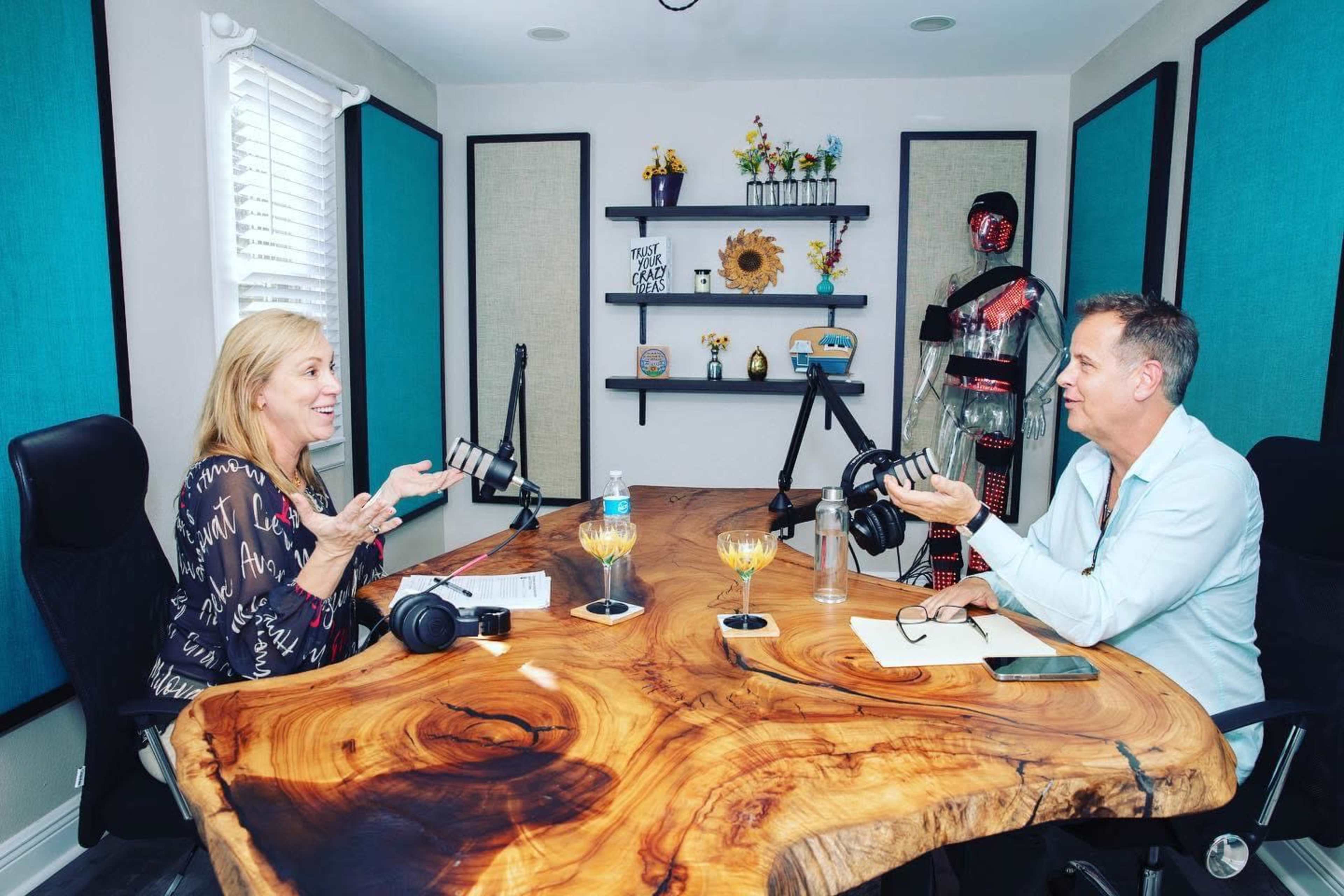 Two people are engaged in conversation at a podcast recording studio with microphones, a large wooden table, and colorful wall decor.