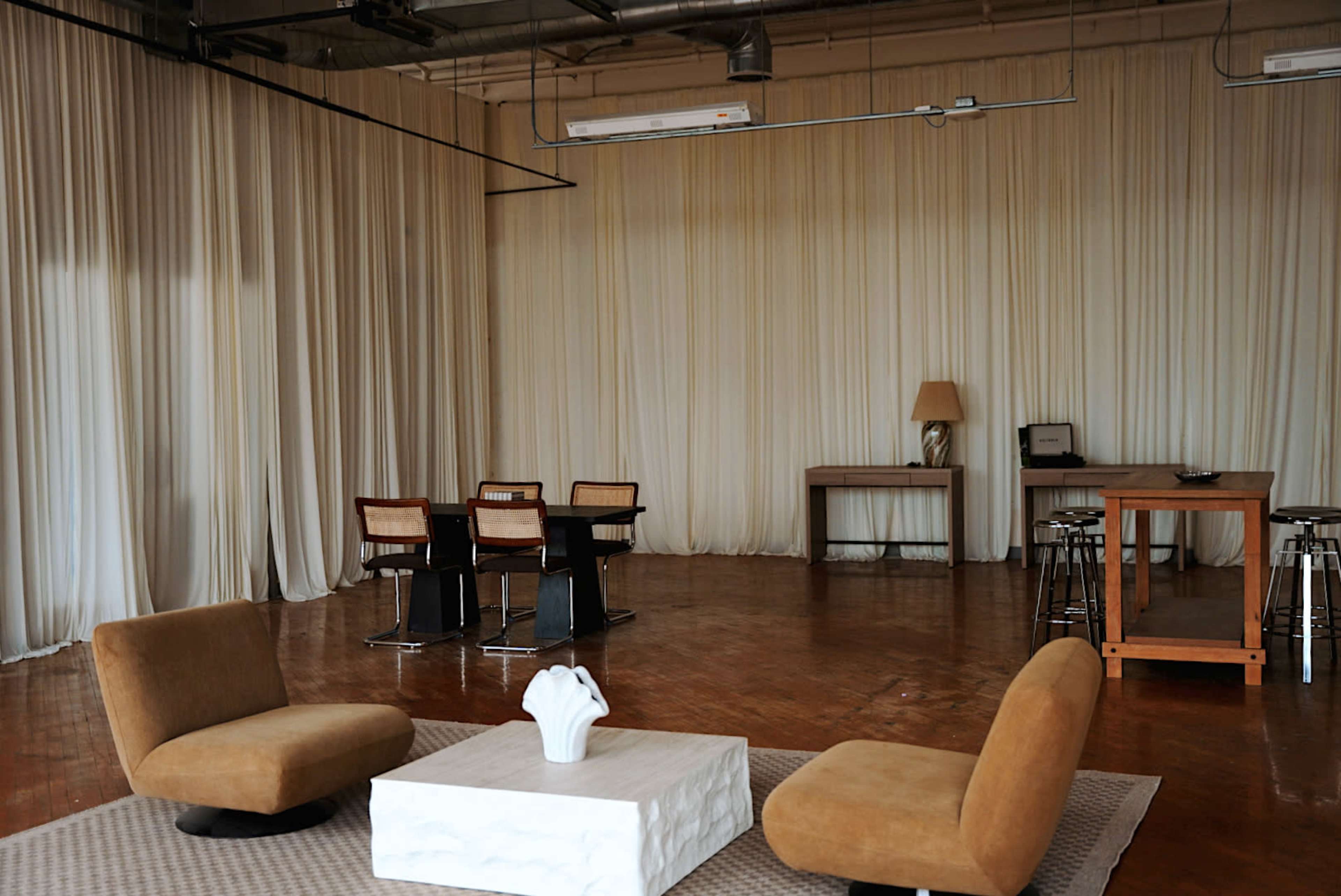 The image shows a spacious, minimalist interior with two brown chairs, a white coffee table, and a dining area with a dark table and chairs under soft, draped curtains.