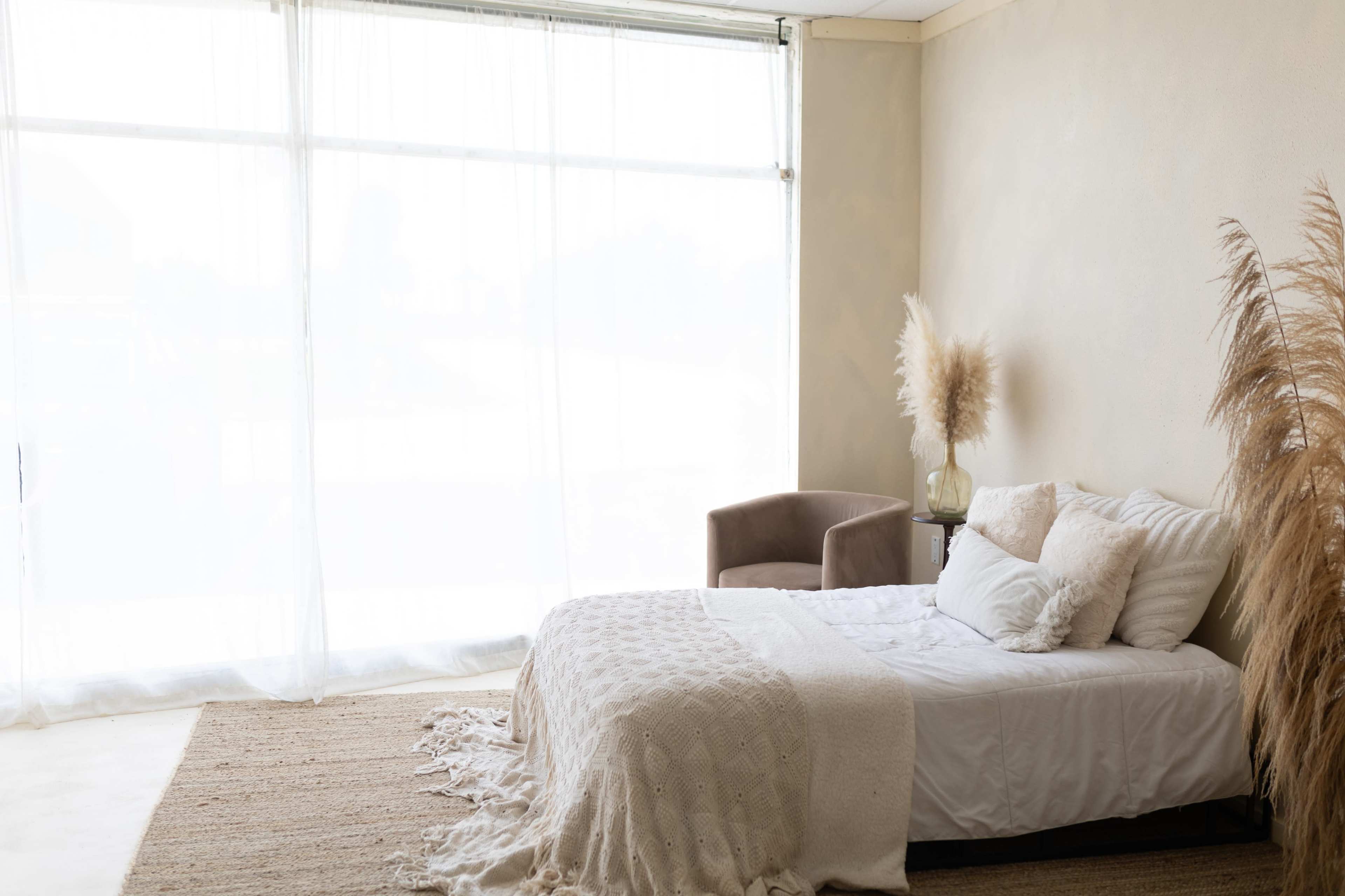 A minimalist bedroom features a bed with a light blanket, a chair, and decorative plants beside large sheer curtains.