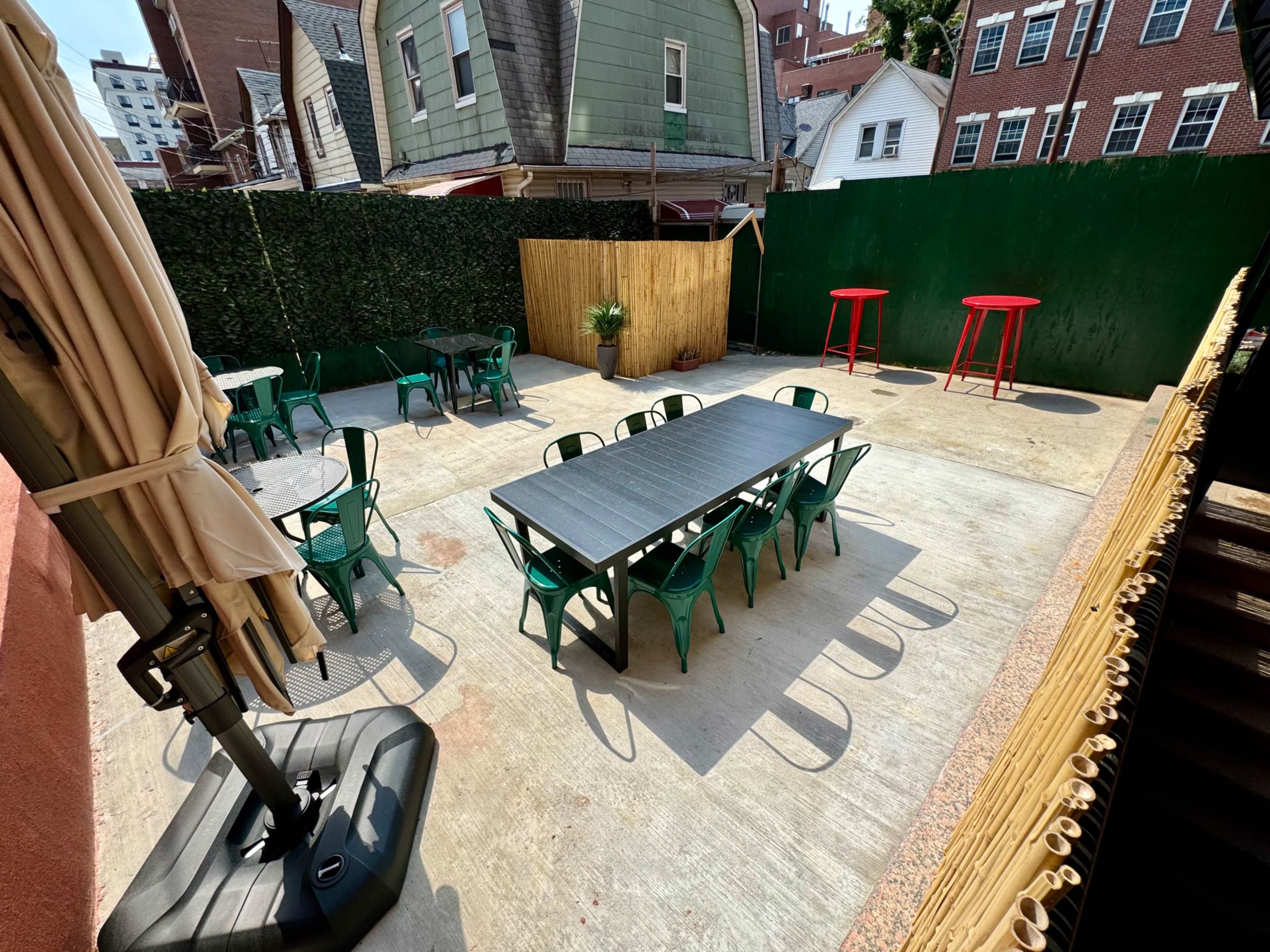The image shows an outdoor patio area with green chairs and tables, flanked by green fencing and red bar tables, set against a backdrop of buildings.