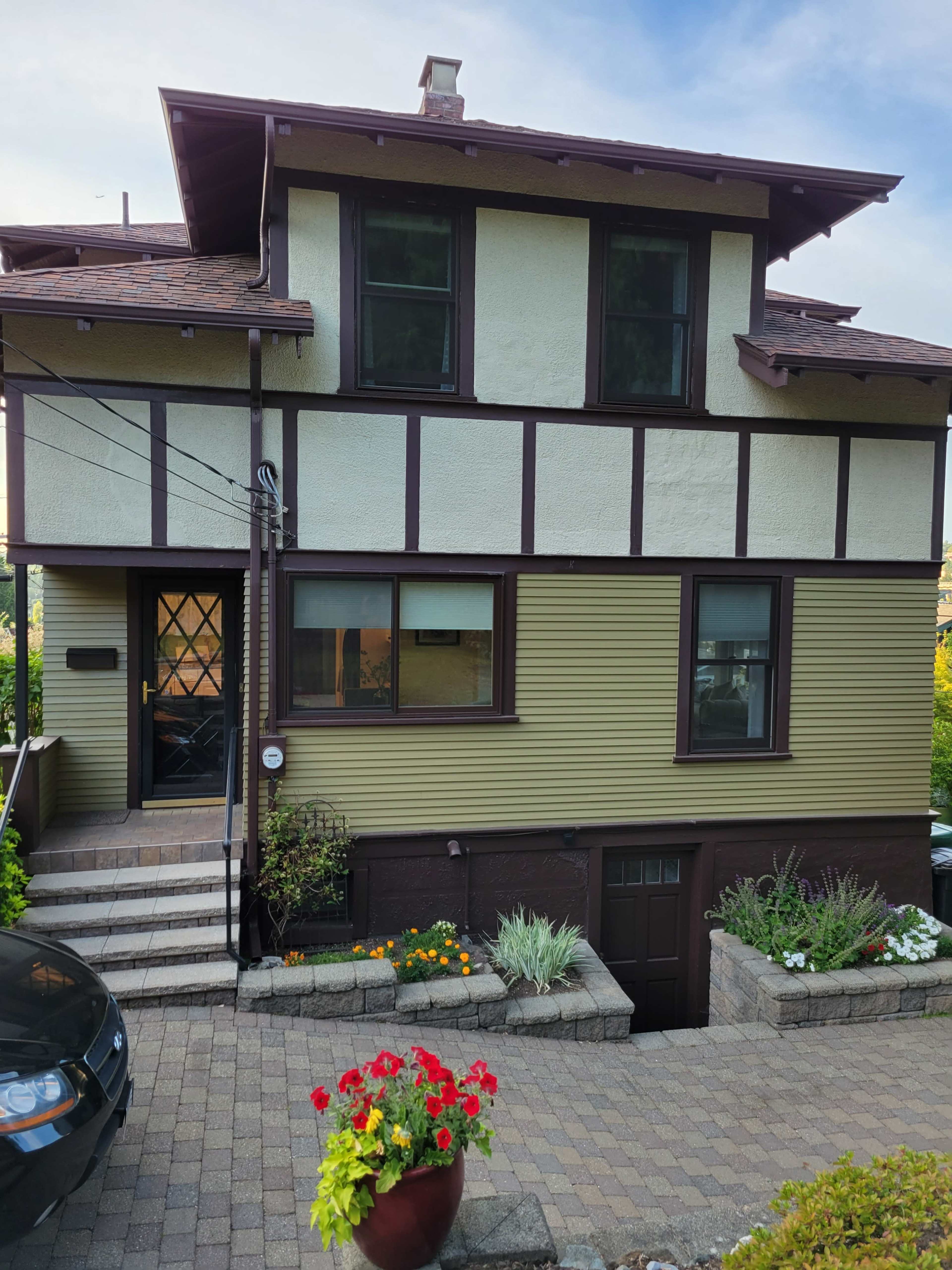 The image shows a two-story house with a combination of yellow and brown exterior, featuring a front entrance with steps and landscaped flower beds.