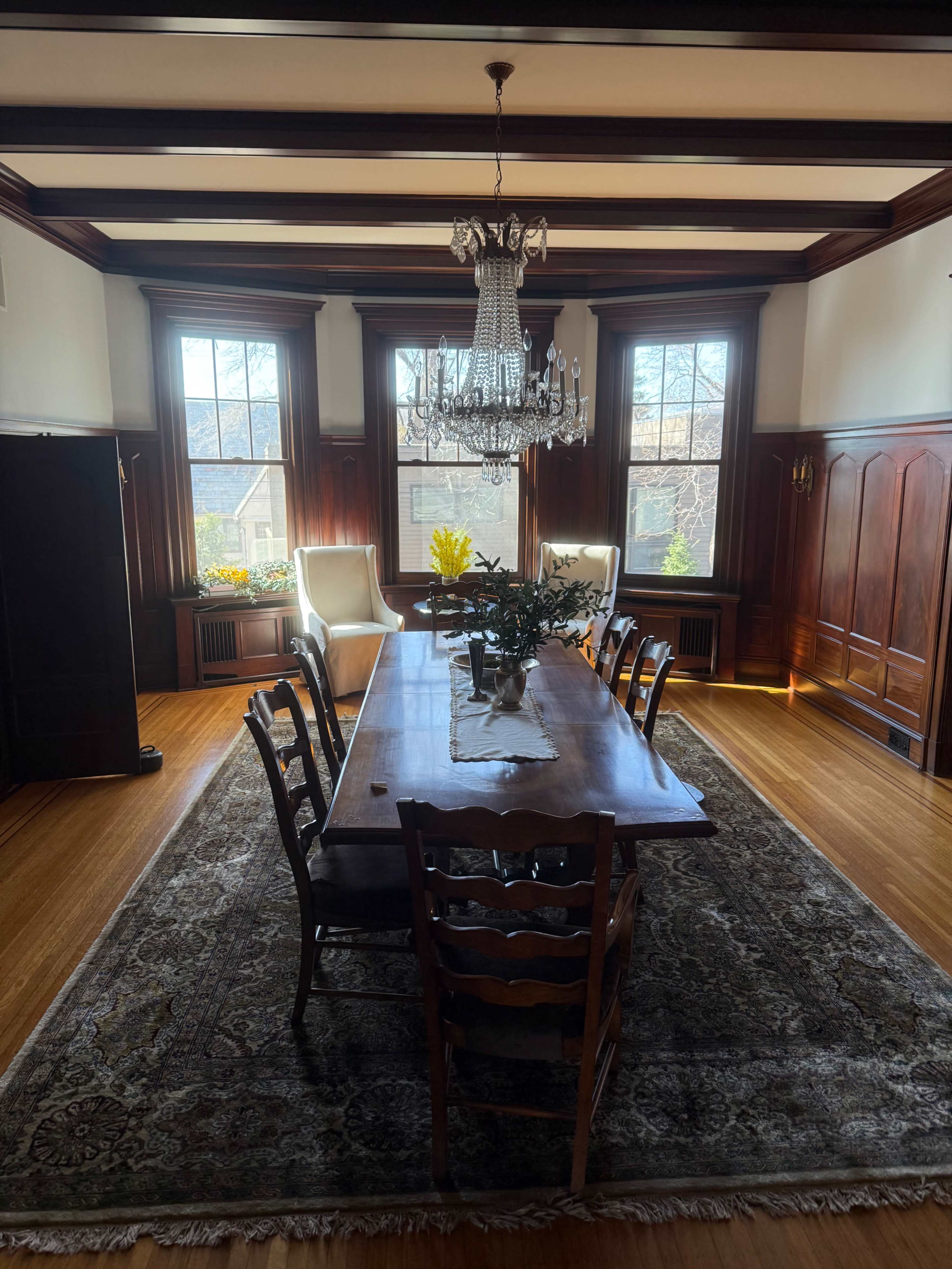 A wooden dining table is set in a well-lit room with large windows, surrounded by chairs, and a chandelier hangs from the ceiling.