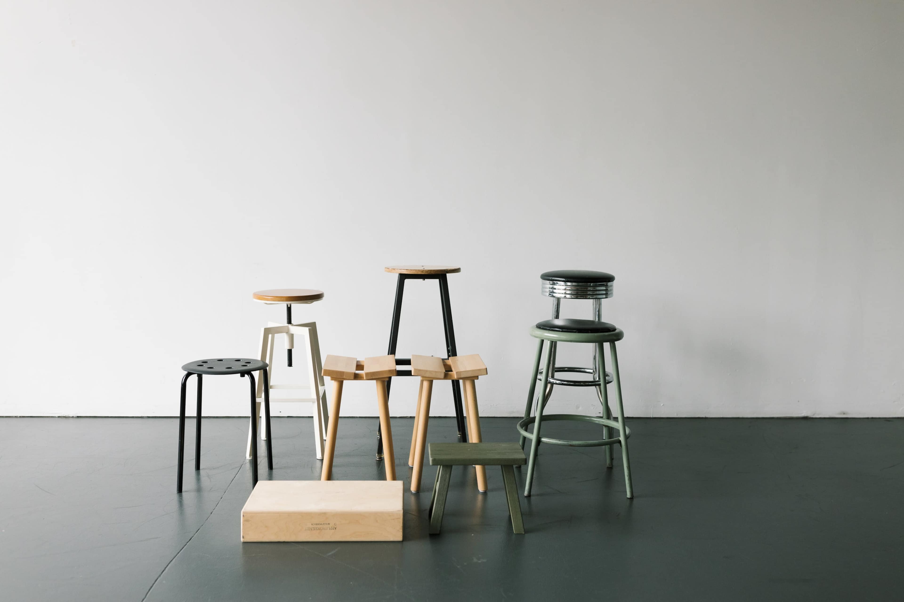 A variety of stools made from different materials and in various heights are arranged on a smooth floor against a plain wall.