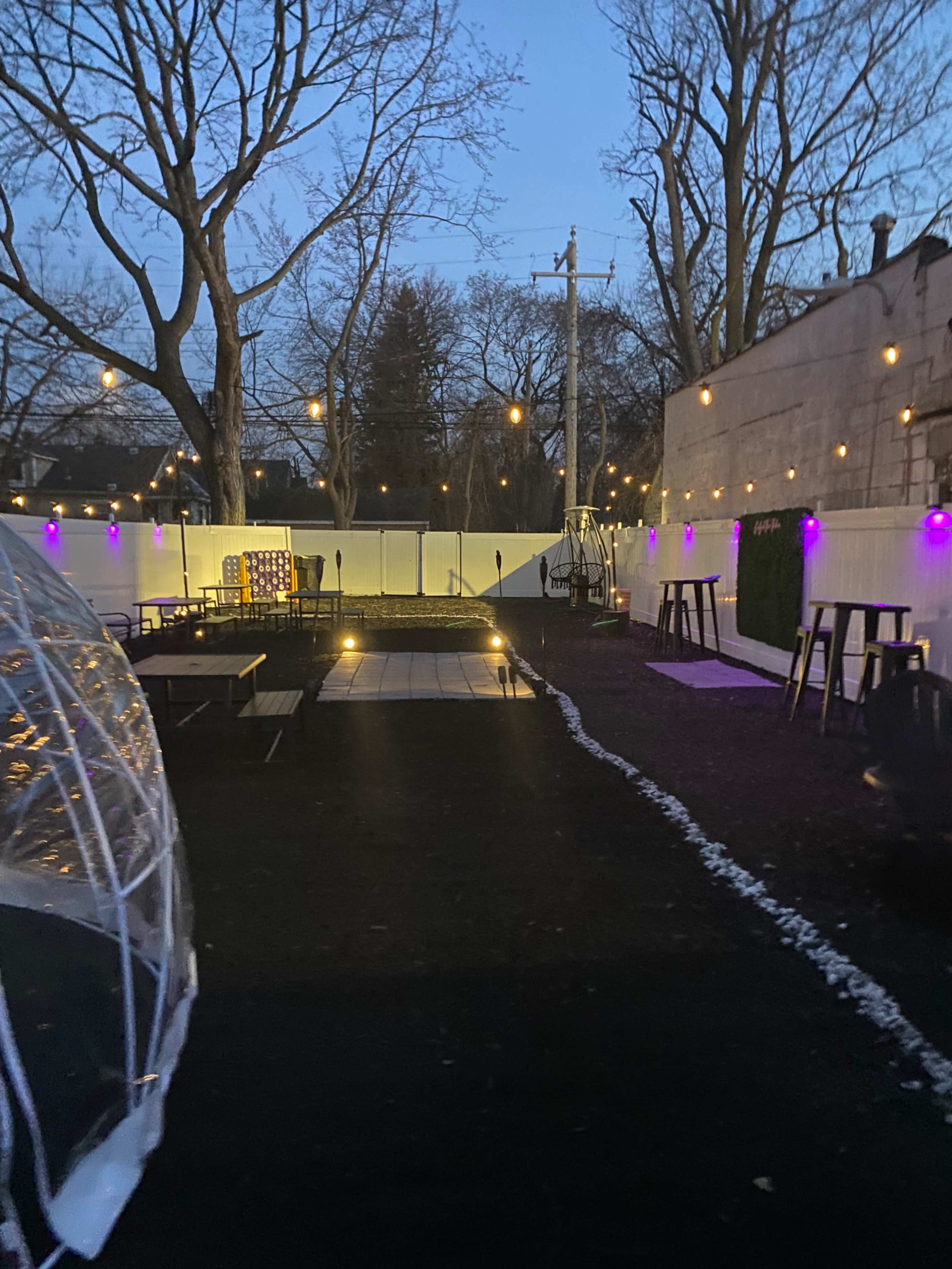 The image shows a landscaped outdoor area with tables, string lights, and a pathway leading through a garden space during dusk.