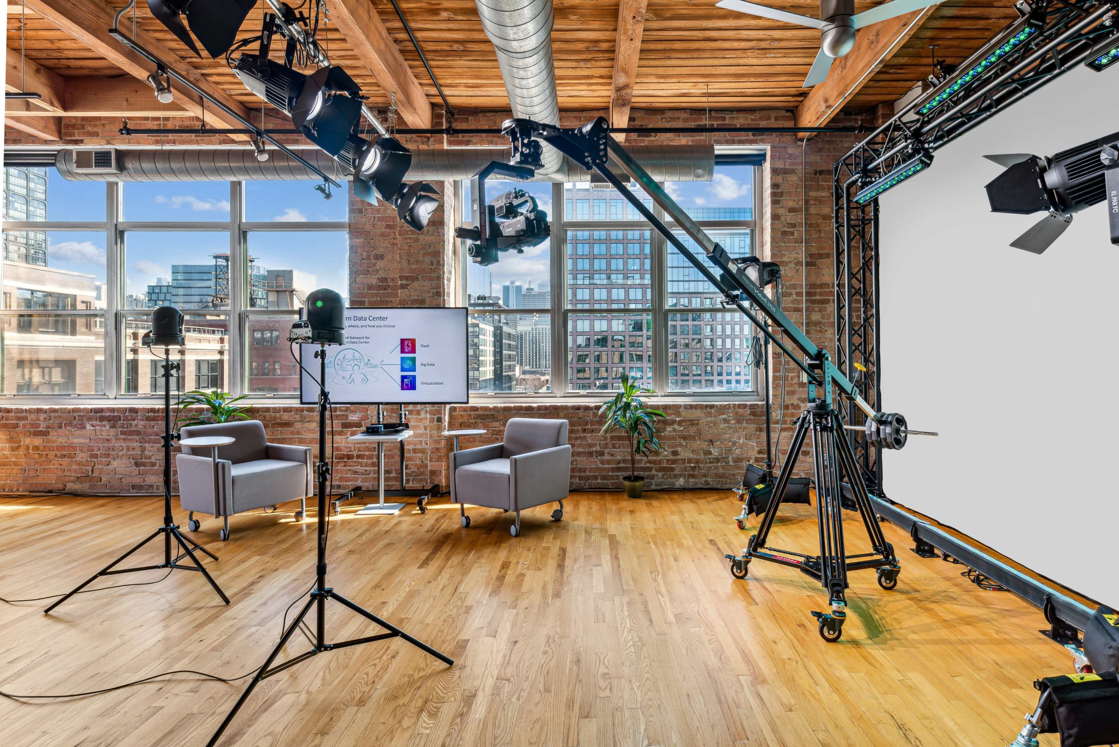A modern studio setup features two chairs, a presentation screen, and professional lighting equipment against a backdrop of large windows and exposed brick walls.