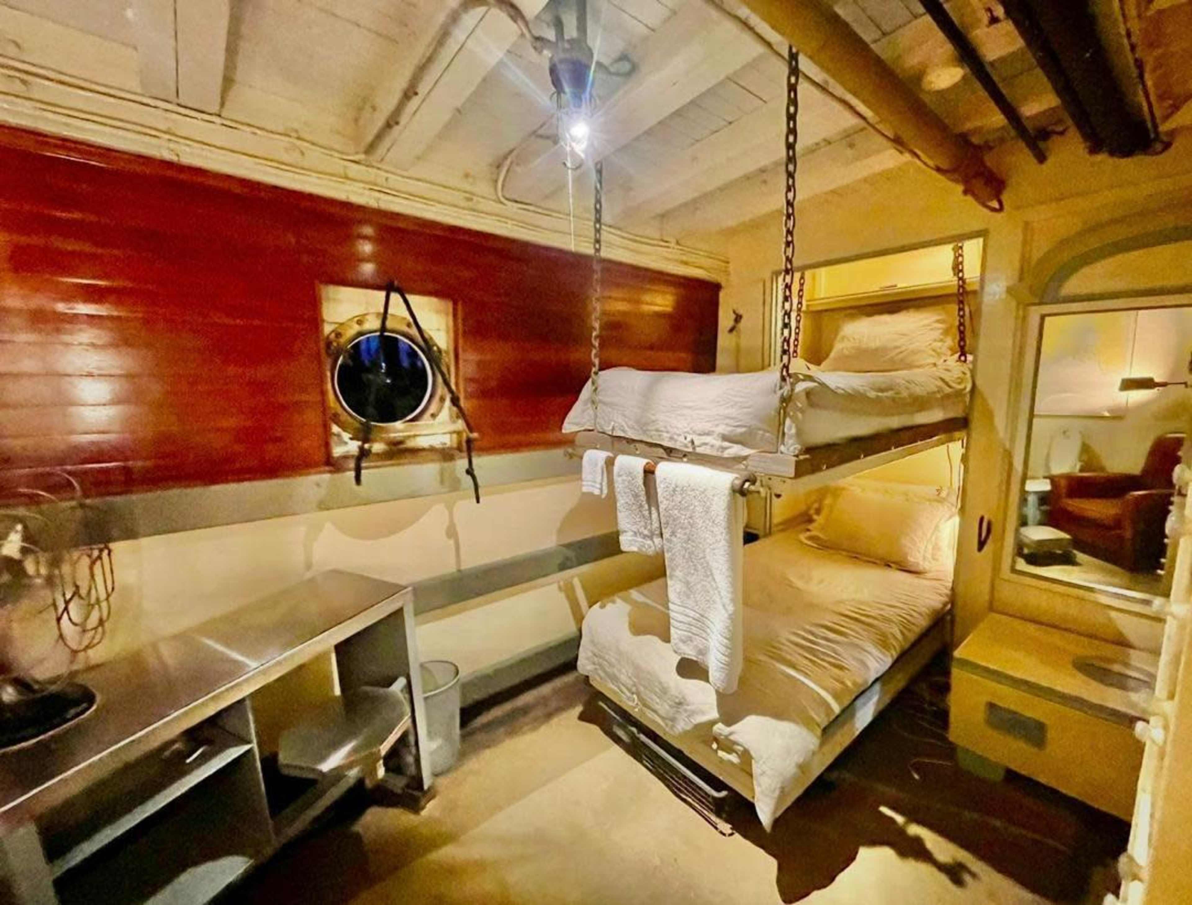 The image shows a small cabin with a bunk bed, a circular window, and a desk.