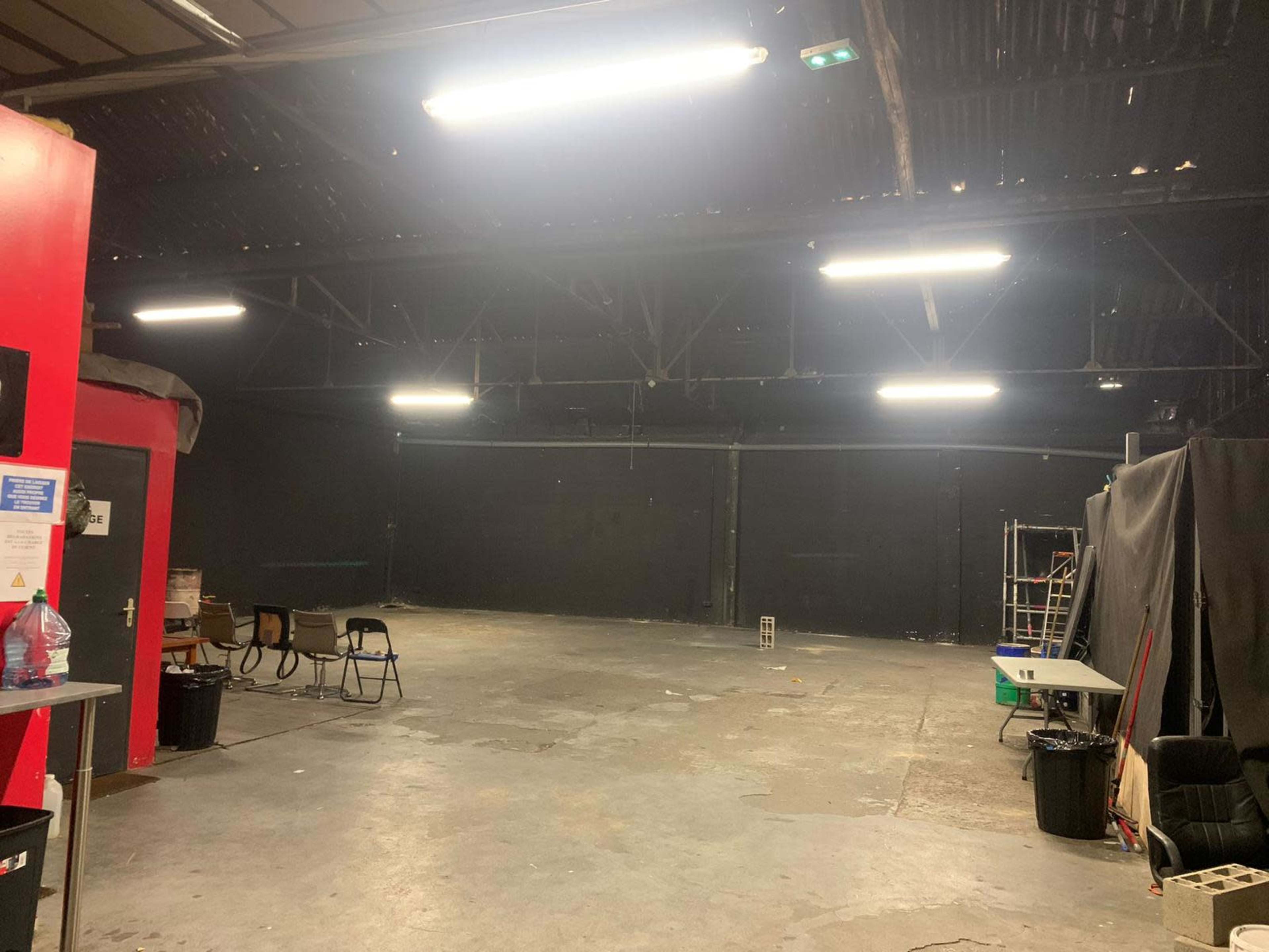 A spacious, dimly lit warehouse interior features black walls, a concrete floor, and various furniture items along the perimeter.