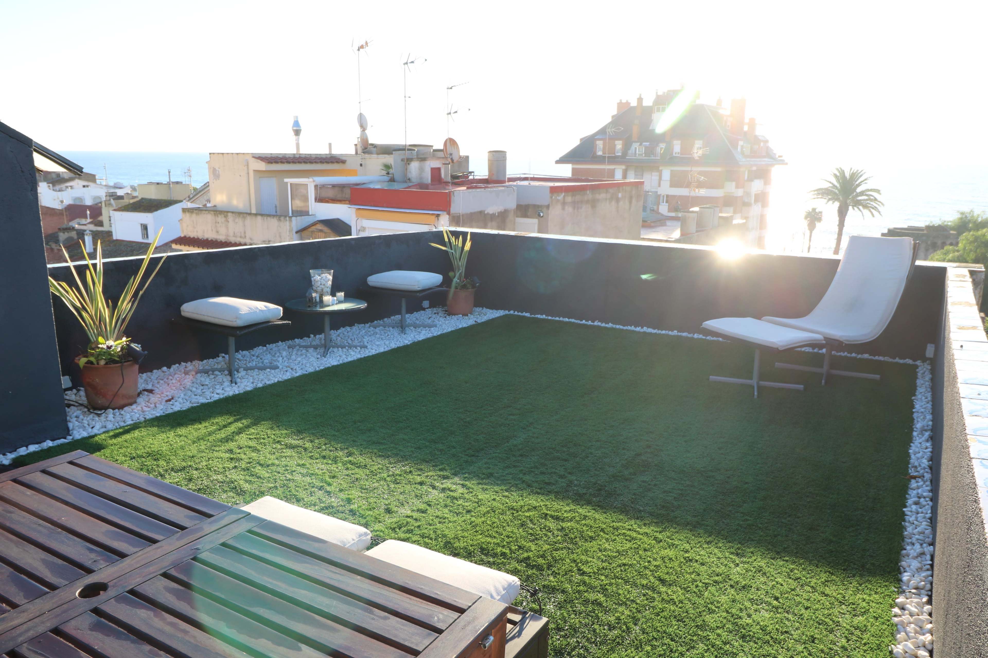 A rooftop terrace features artificial grass, a wooden table, lounge chairs, and sea views.