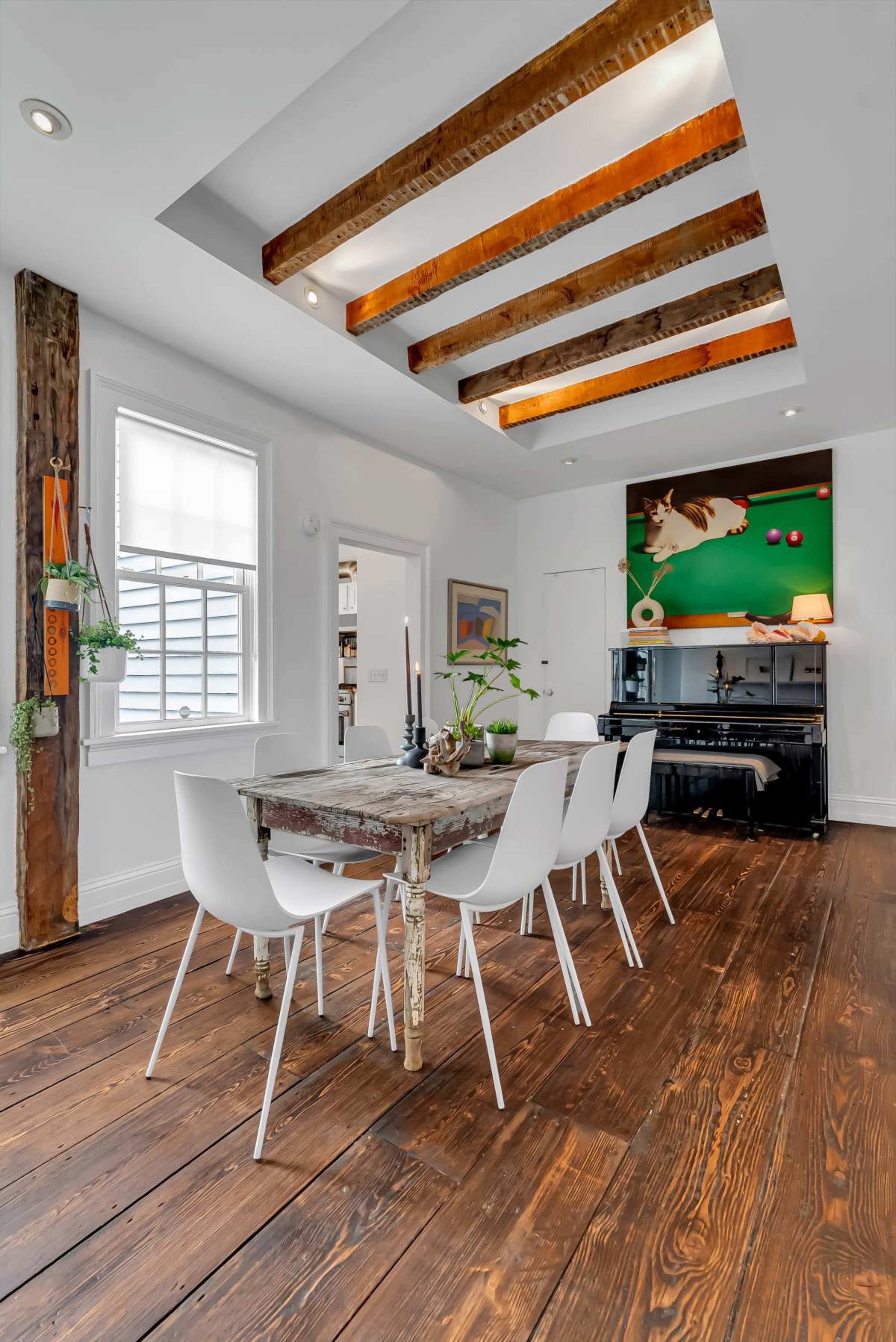 The image shows a dining area with a wooden table and six white chairs beneath a ceiling with exposed beams, while a green wall with artwork and a black piano are in the background.