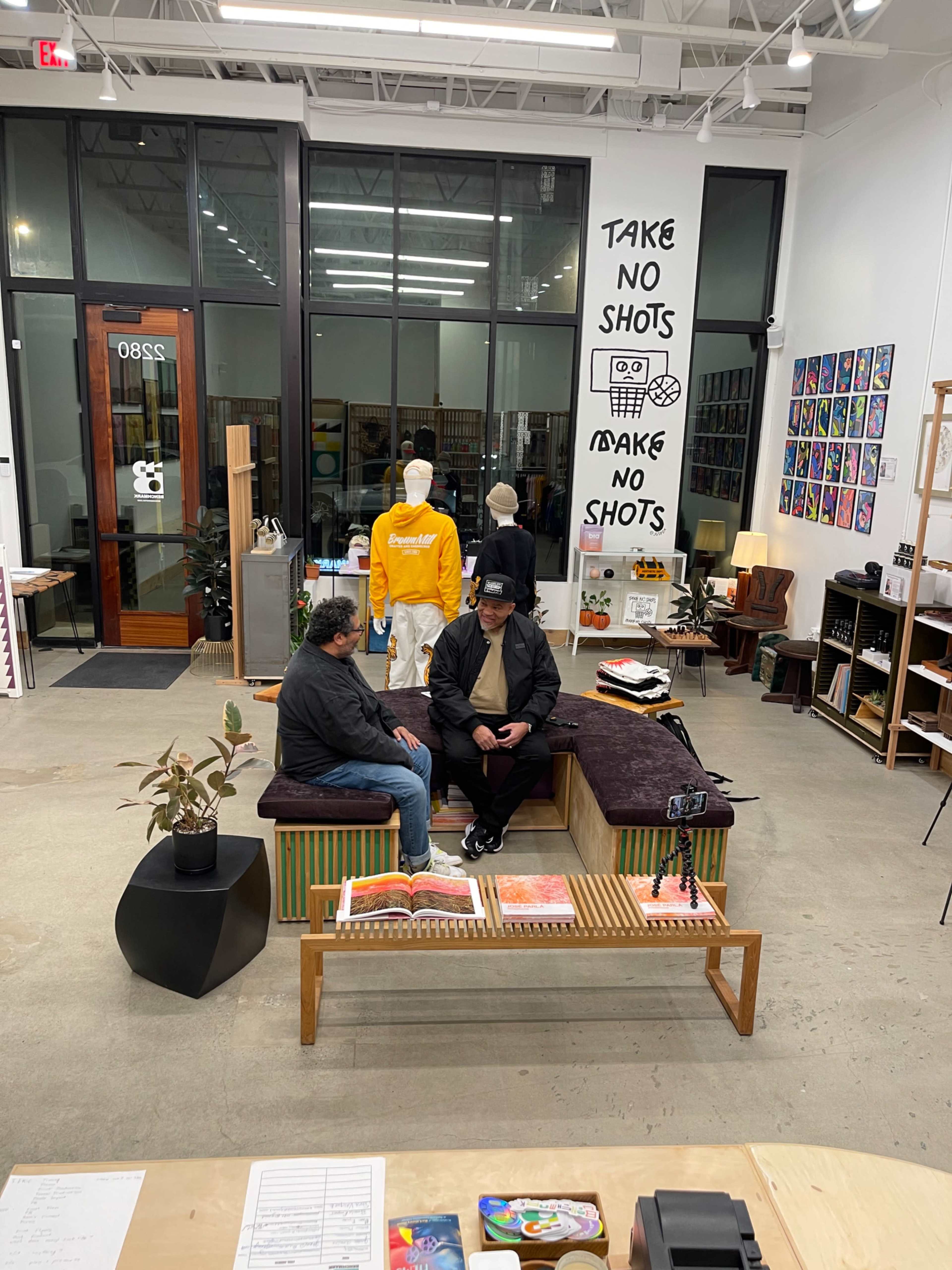 Two men are seated on a bench in a modern shop with furniture and decor displayed around them.