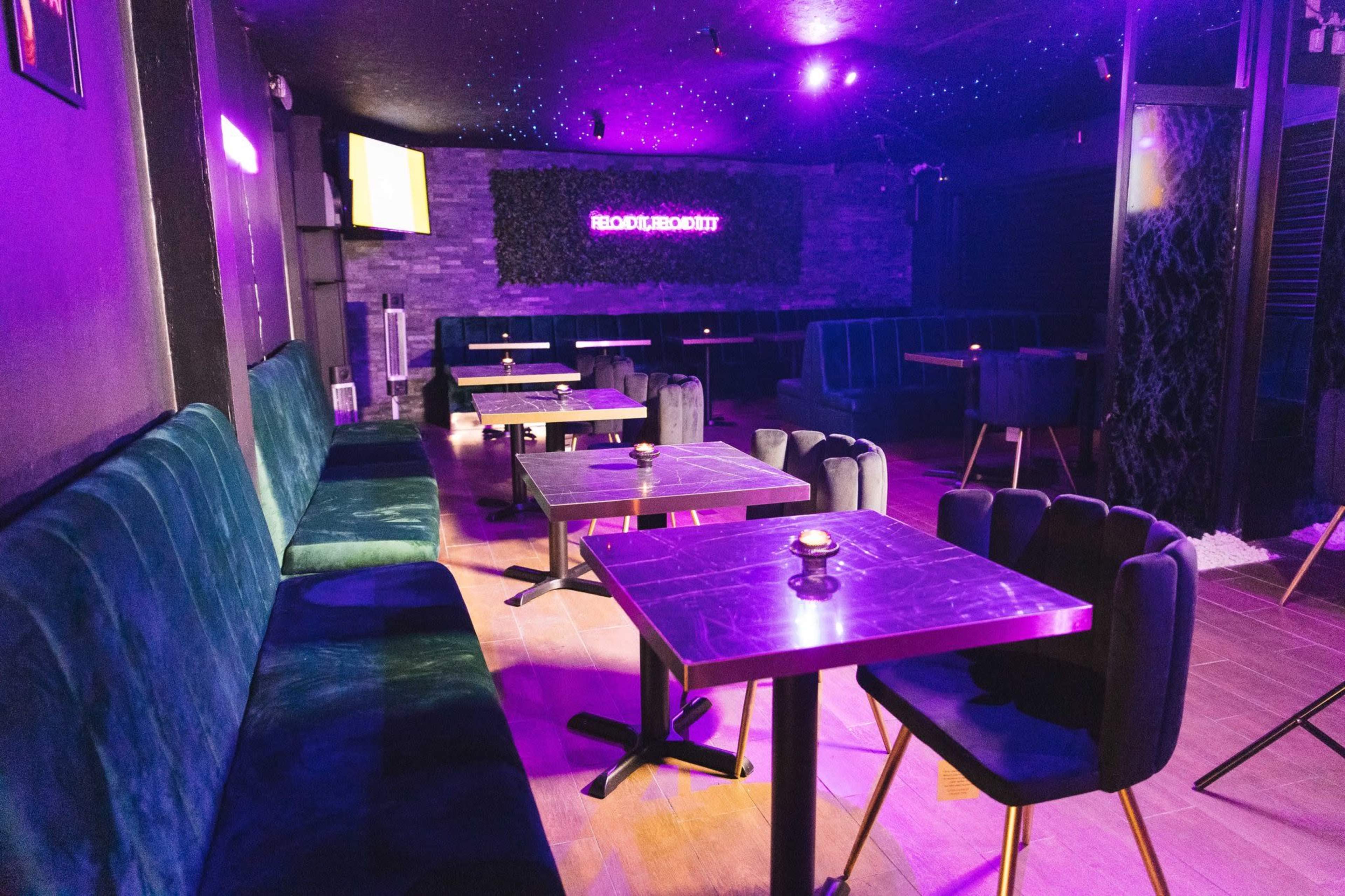 The image shows a dimly lit lounge or club interior featuring black and teal seating, small tables, and purple lighting.