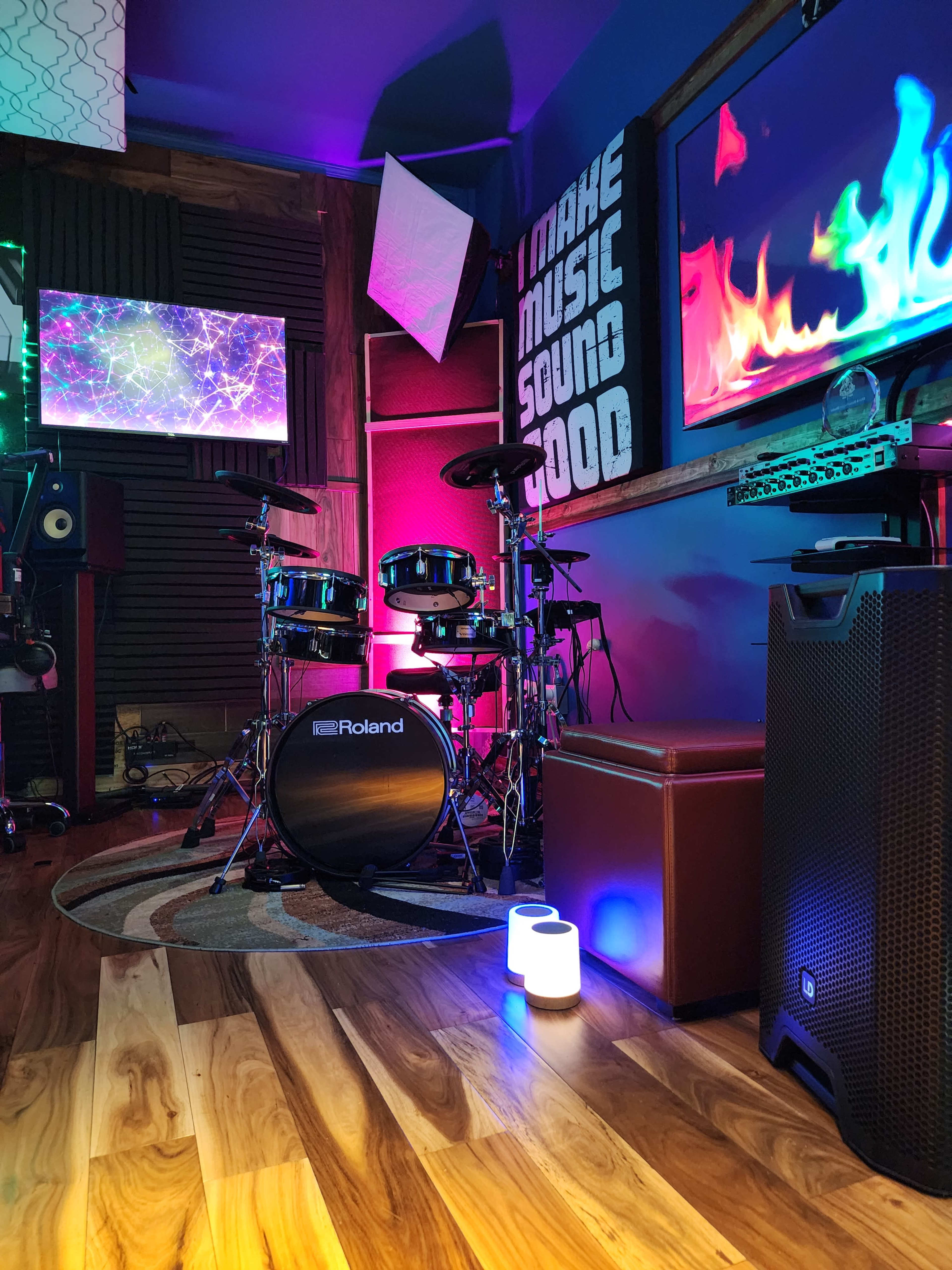 A music studio setup with electronic drums, colorful lighting, and various audio equipment arranged in a cozy space.